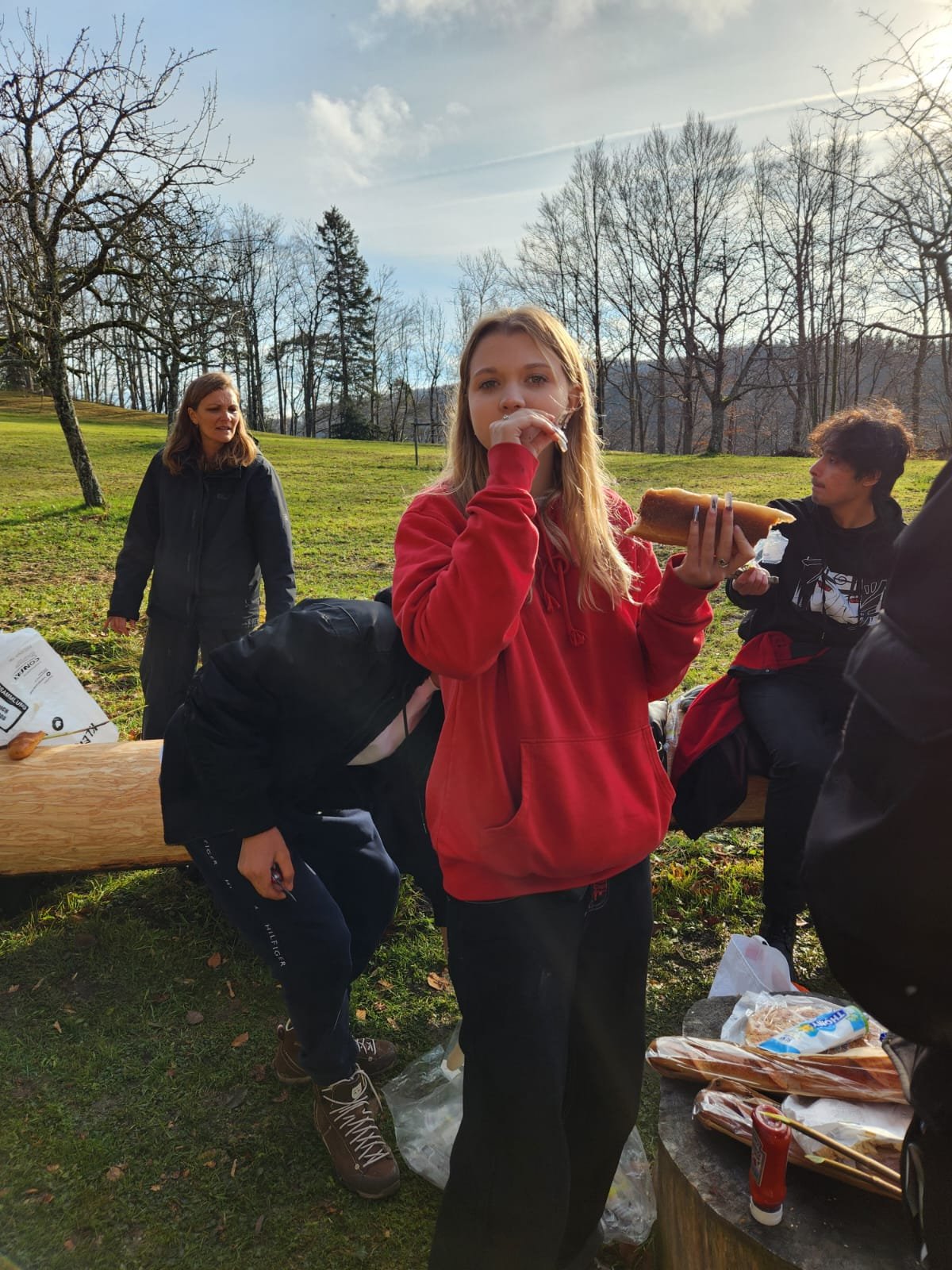 Gruppe von Menschen im Freien, eine Person isst einen Hotdog, umgeben von Bäumen und Natur, mit Picknickzubehör auf einem Tisch.