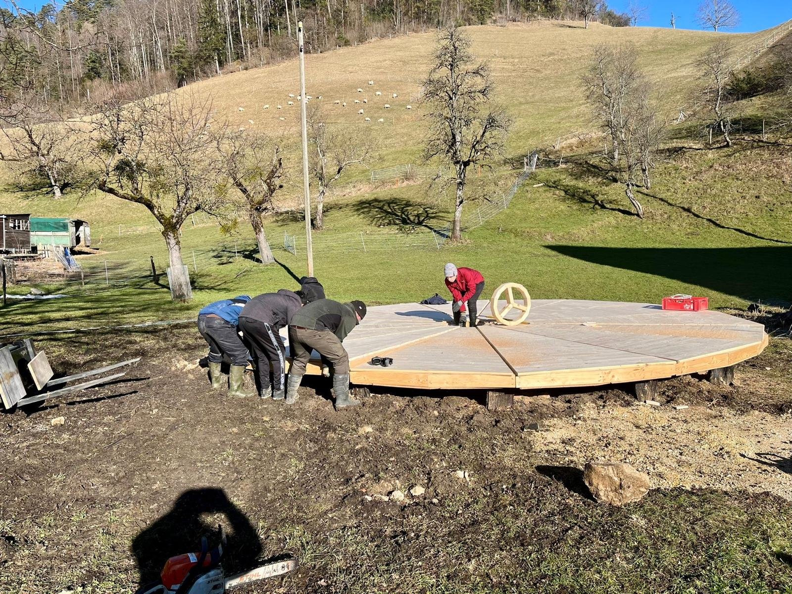 Mehrere Personen arbeiten auf einem Feld an einer kreisförmigen Holzkonstruktion. Die Umgebung ist ländlich mit Bäumen, einem Hügel und einem kleinen Gebäude im Hintergrund. Eine Person steht in der Mitte, während die anderen am Rand arbeiten. Es ist
