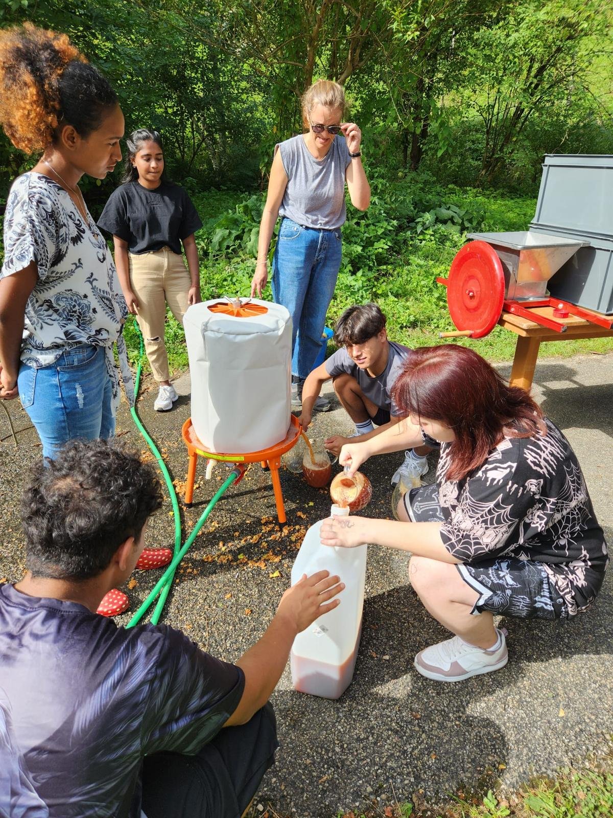 Eine Gruppe von sechs Personen steht im Freien um eine Saftpresse. Sie füllen den ausgepressten Saft in einen großen weißen Behälter. Im Hintergrund ist viel Grün und ein weiteres Gerät zu sehen. Der Tag scheint sonnig zu sein.