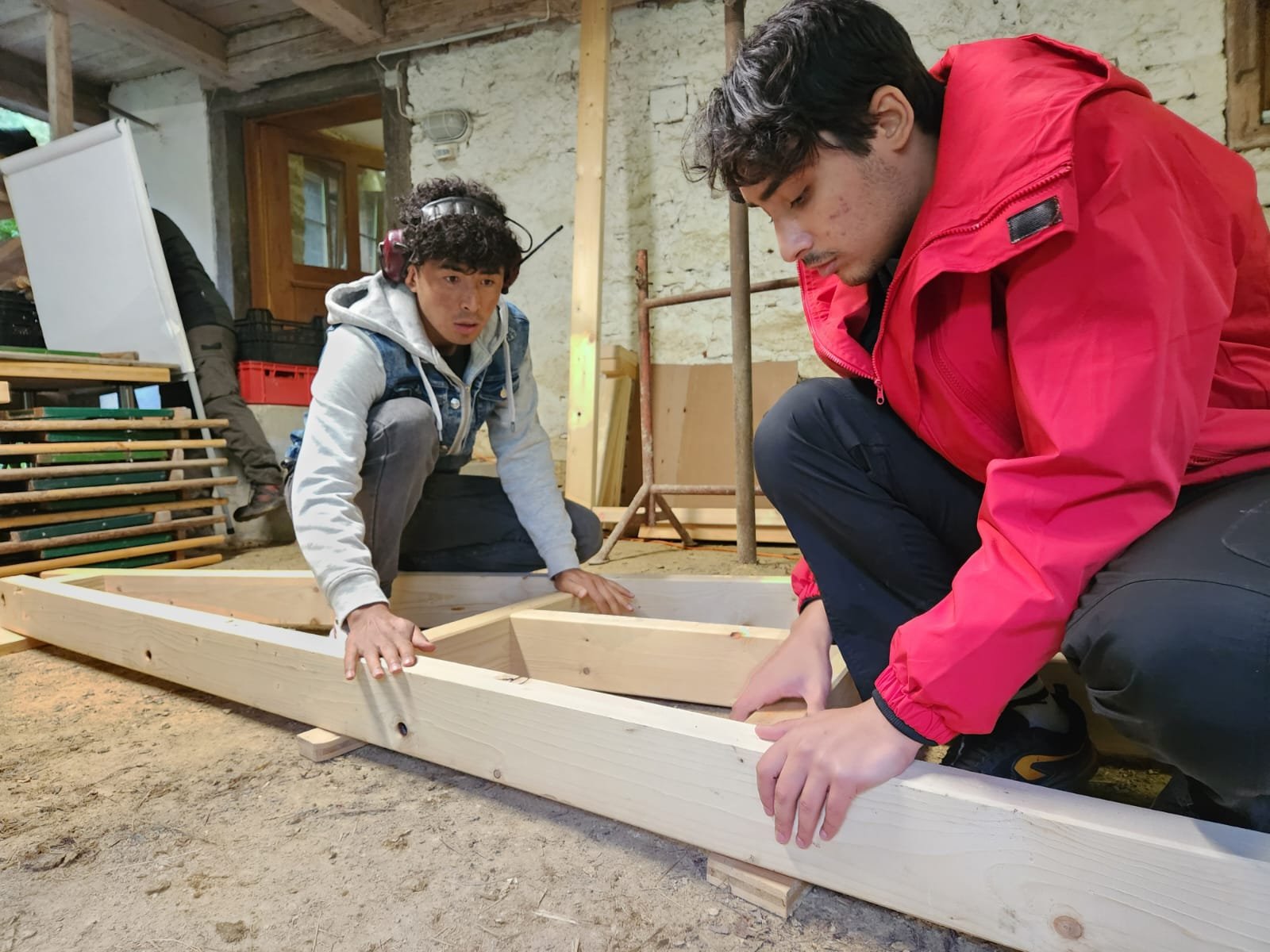 Zwei Personen arbeiten an einem Holzrahmen; eine Person trägt Kopfhörer, die andere eine rote Jacke.