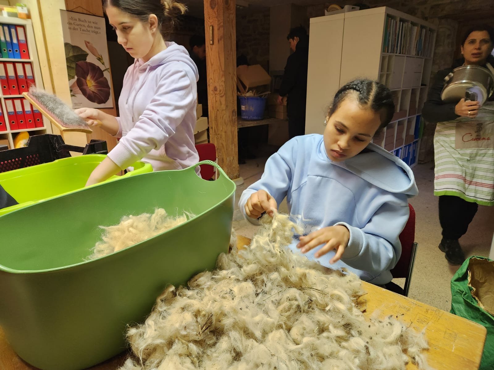 Zwei Personen arbeiten in einem Raum mit Wolle. Eine Person bürstet Wolle über einer großen grünen Plastikwanne, während die andere Person Wolle auf einem Tisch bearbeitet. Im Hintergrund ist eine weitere Person mit einem Topf zu sehen. Der Raum enth