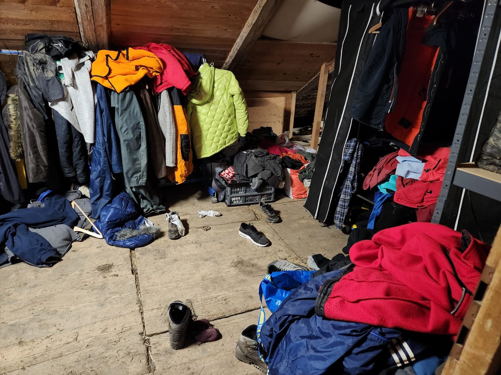 Unordentlicher Raum mit vielen Kleidungsstücken, darunter bunte Jacken, Pullover und Schuhe auf dem Boden und Kleiderständern.