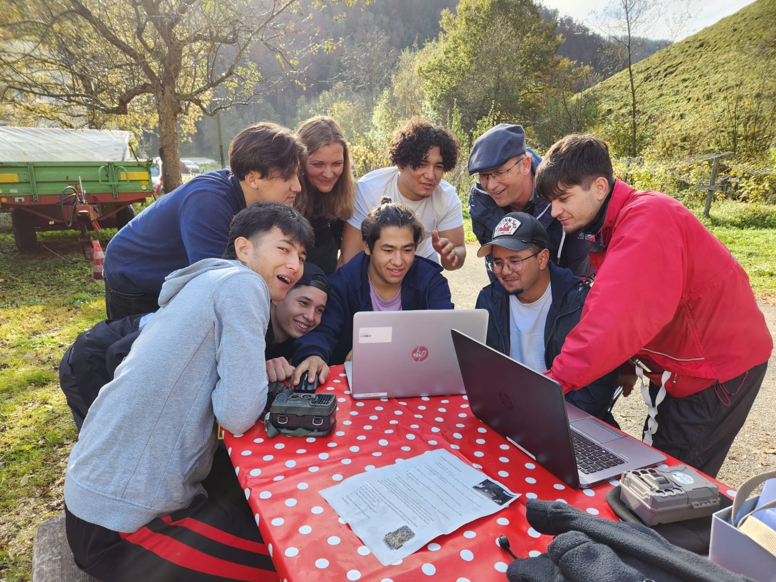 Eine Gruppe von neun Personen schaut gemeinsam auf zwei Laptops im Freien. Sie sitzen an einem Tisch mit rot-weiß gepunkteter Tischdecke. Im Hintergrund ist eine grüne Wiese und ein Anhänger zu sehen. Einige Zettel liegen auf dem Tisch.