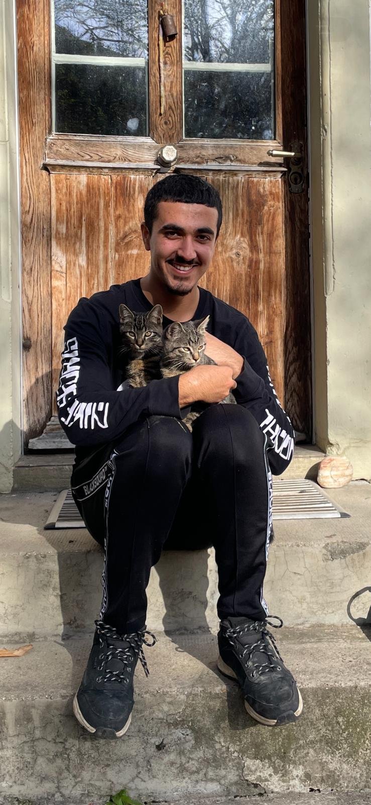 Eine Person sitzt vor einer Holztür und hält zwei Katzen im Arm. Die Person trägt schwarze Sportkleidung und lächelt. Die Umgebung ist außen auf einer Treppe.