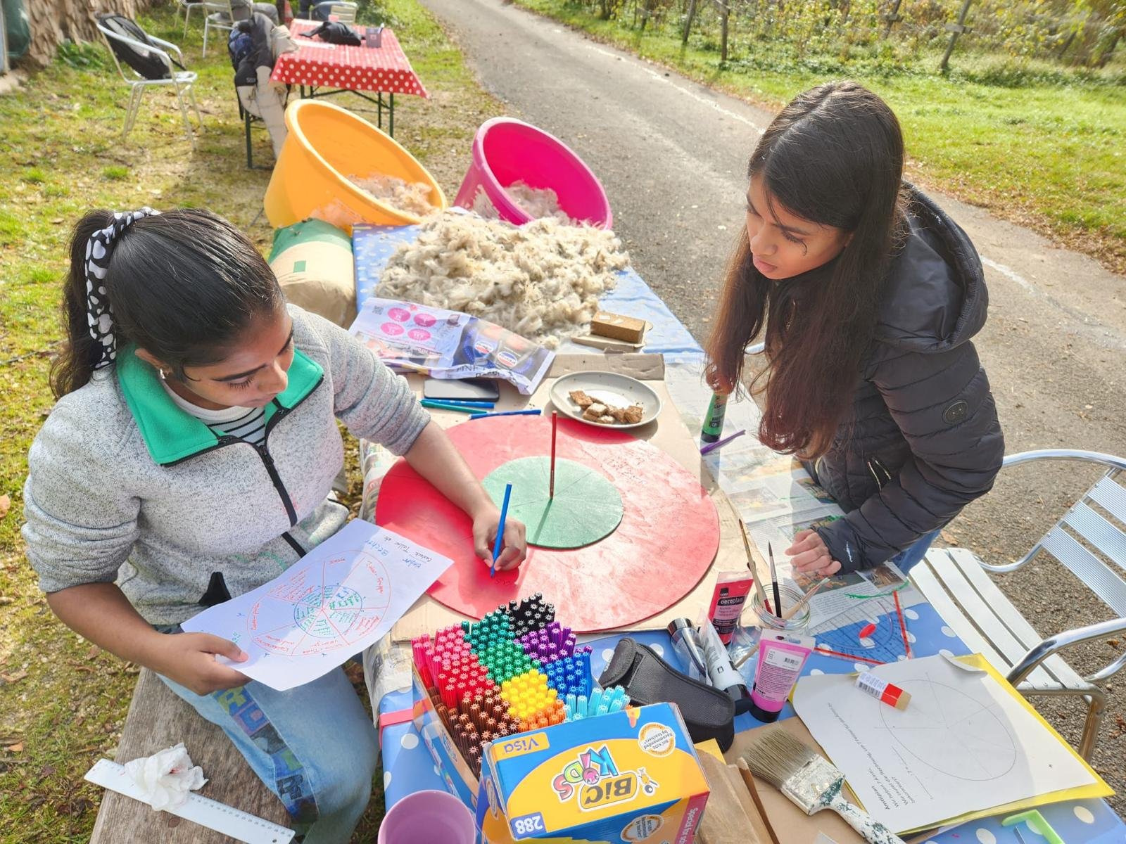 Zwei junge Menschen sitzen an einem Tisch im Freien und arbeiten an einem Kunst- oder Bastelprojekt. Auf dem Tisch liegen farbige Stifte, Papier und Malutensilien. Im Hintergrund sind bunte Behälter mit Materialien zu sehen.