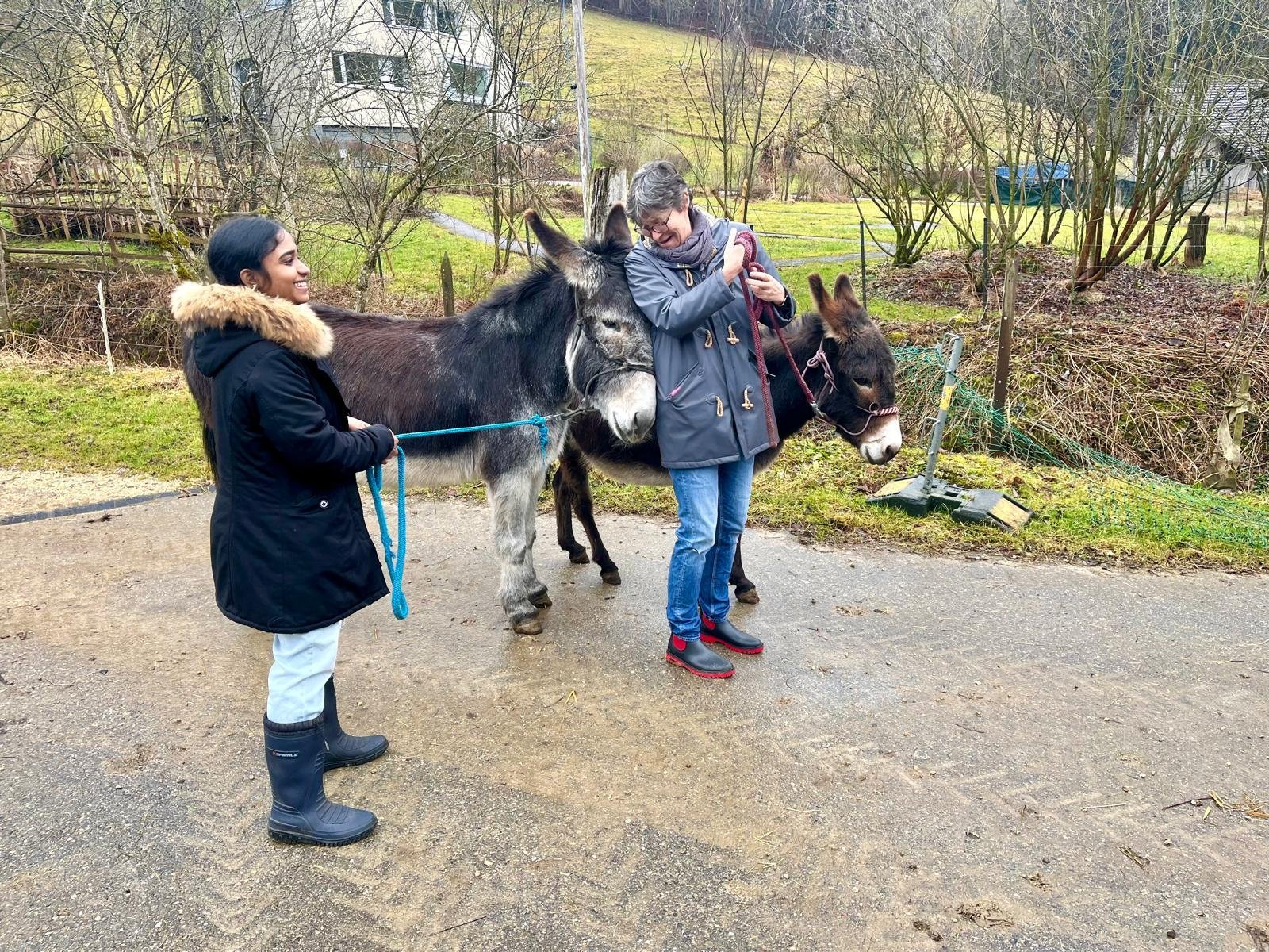 Zwei Personen spazieren mit zwei Eseln an Leinen auf einem ländlichen Weg. Eine Person trägt eine schwarze Jacke mit Fellkragen, die andere eine graue Jacke. Im Hintergrund sind Bäume und ein Haus zu sehen.