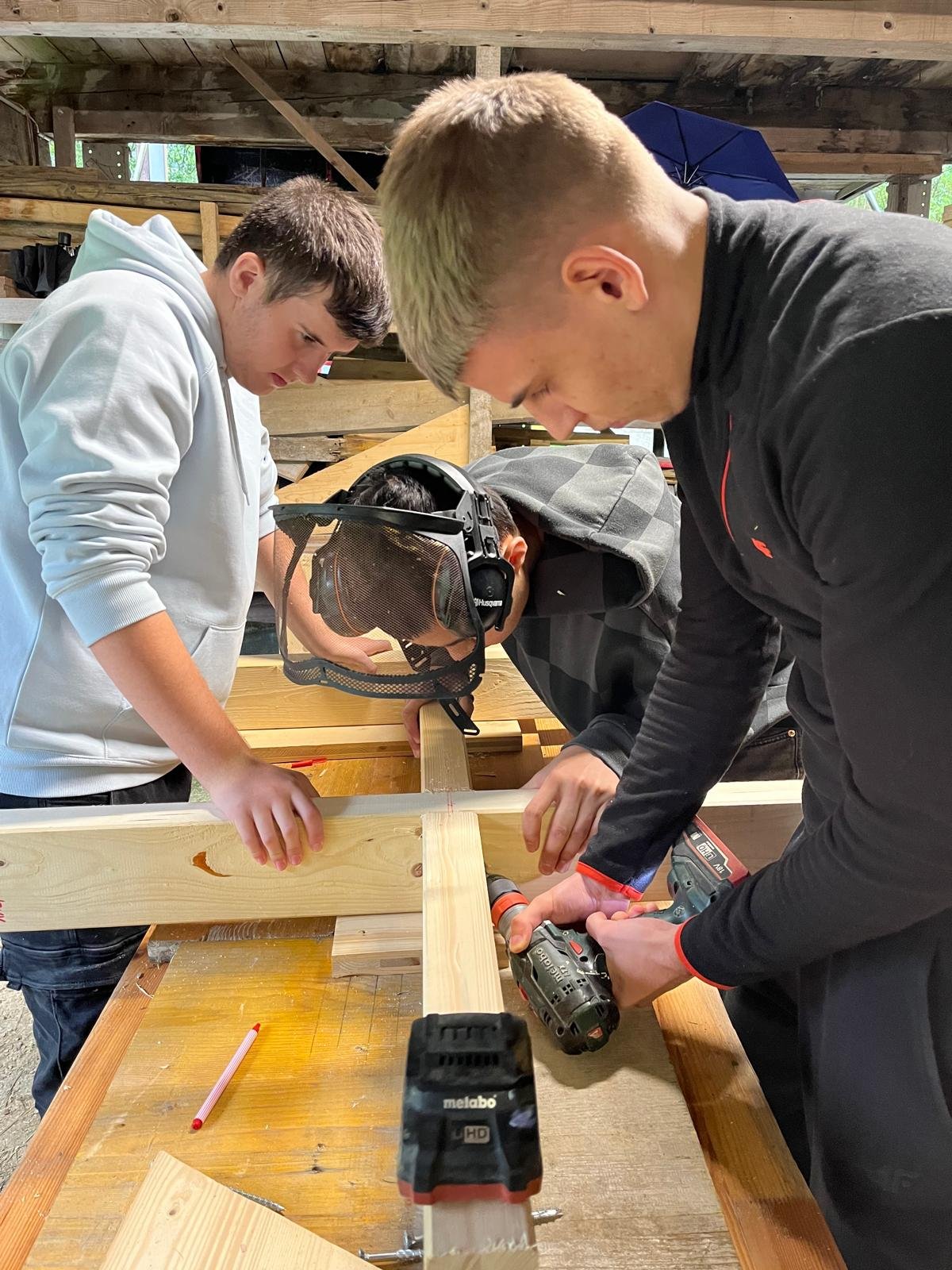 Drei Jugendliche arbeiten konzentriert an einem Holzprojekt in einer Werkstatt. Einer benutzt einen Akkuschrauber, während die anderen das Holz stabilisieren. Schutzkleidung und Werkzeuge sind zu sehen.