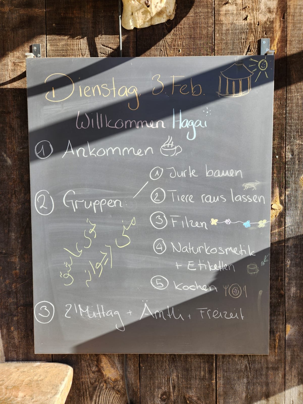 Tafel mit einer Tagesordnung für Dienstag, den 3. Februar. Oben steht "Willkommen Hagai", darunter sind Punkte wie Ankommen, Gruppenaktivitäten wie Jurte bauen, Tiere rauslassen, Filzen, Naturkosmetik und Etiketten erstellen sowie Kochen. Unten ist d
