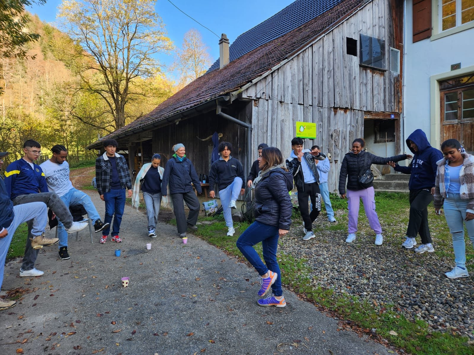 Eine Gruppe von jungen Leuten steht draußen auf einem Weg vor einem alten Holzgebäude. Sie scheinen an einer Art Gruppenspiel teilzunehmen, bei dem einige Personen ein Bein anheben. Der Hintergrund zeigt Bäume und blauen Himmel, was auf einen klaren