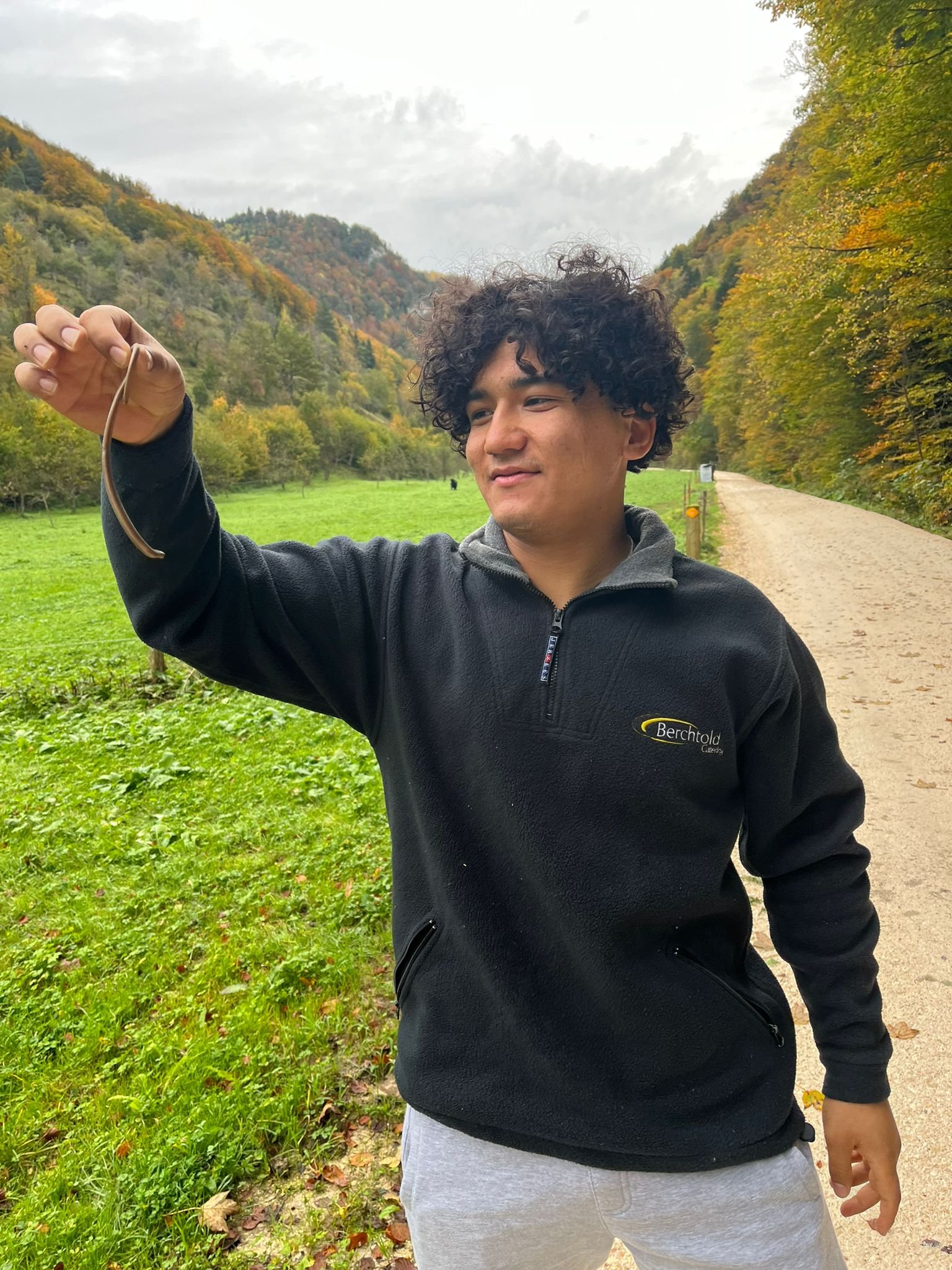 Ein junger Mann steht auf einem Waldweg und hält einen Wurm oder kleinen Ast in der Hand. Im Hintergrund ist eine herbstliche Landschaft mit Bäumen und Hügeln zu sehen.