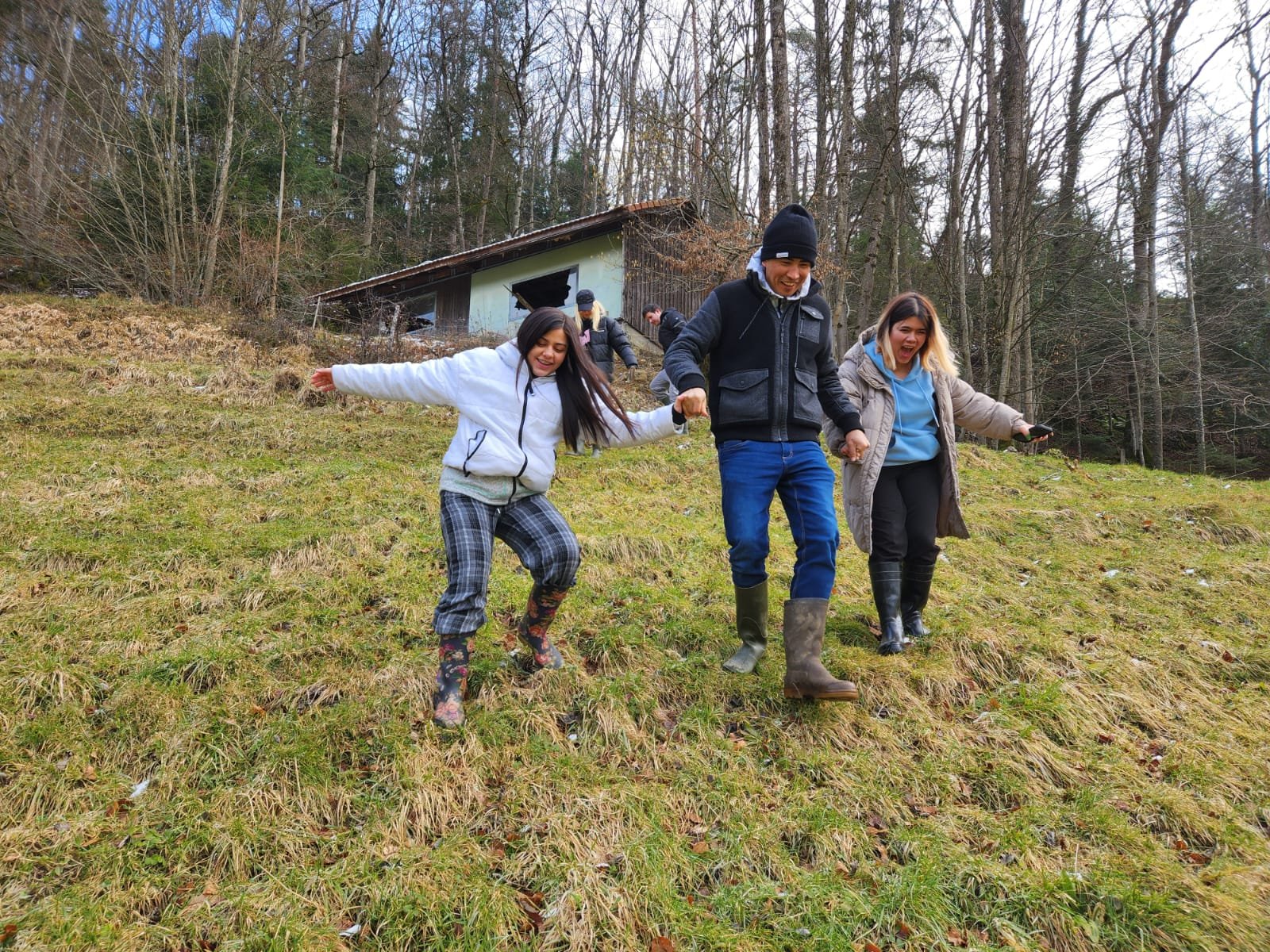 Drei Personen auf einem grasbewachsenen Hügel, die Spaß haben und den Abhang hinunterlaufen. Im Hintergrund ist eine kleine Hütte und Bäume zu sehen.