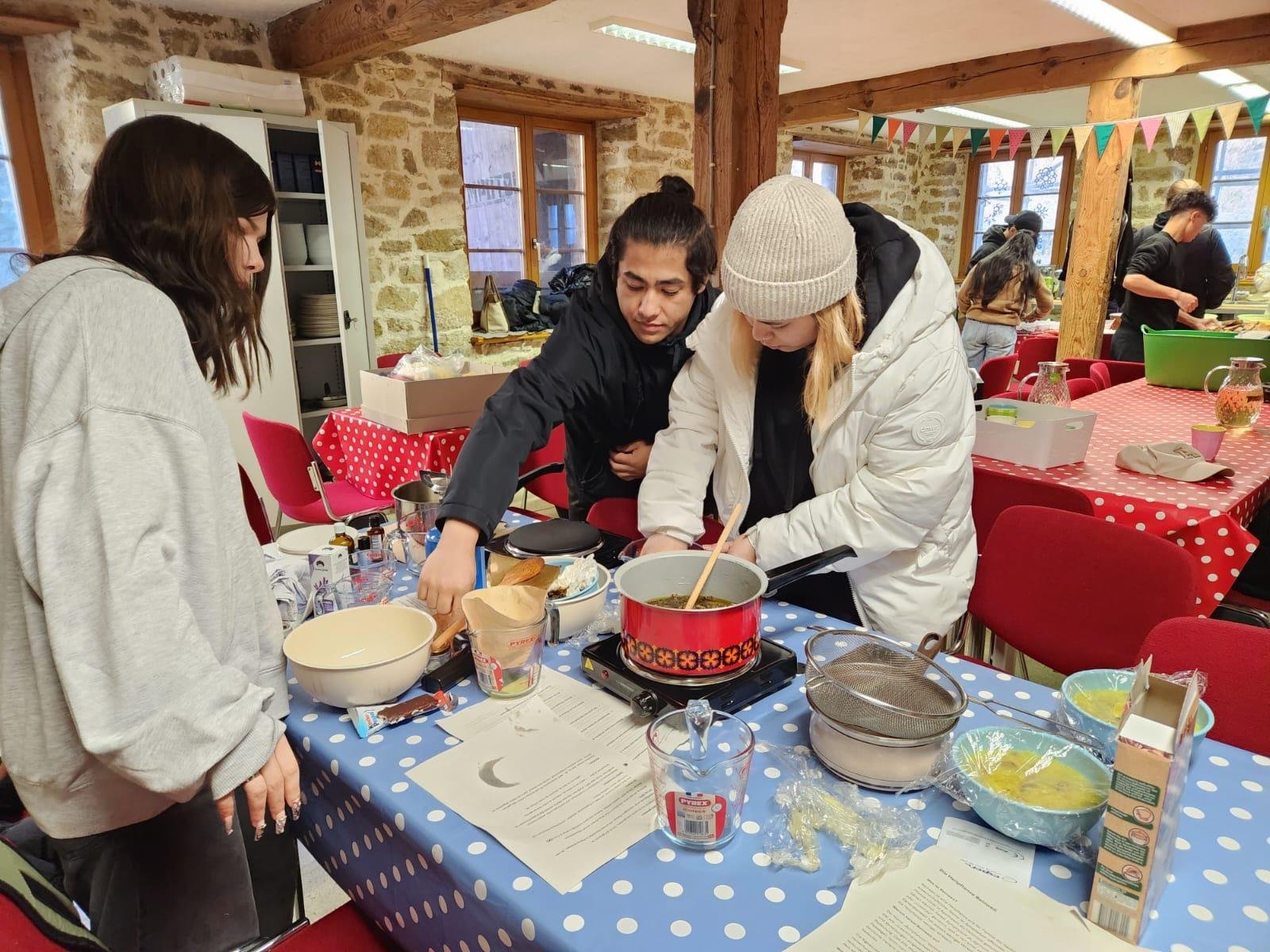 Personen kochen in einer Küche mit Holzbalken. Sie verwenden verschiedene Utensilien und Zutaten, um ein Gericht zuzubereiten. Der Raum ist mit bunten Fähnchen dekoriert, und Tische sind mit gepunkteten Tischdecken bedeckt.