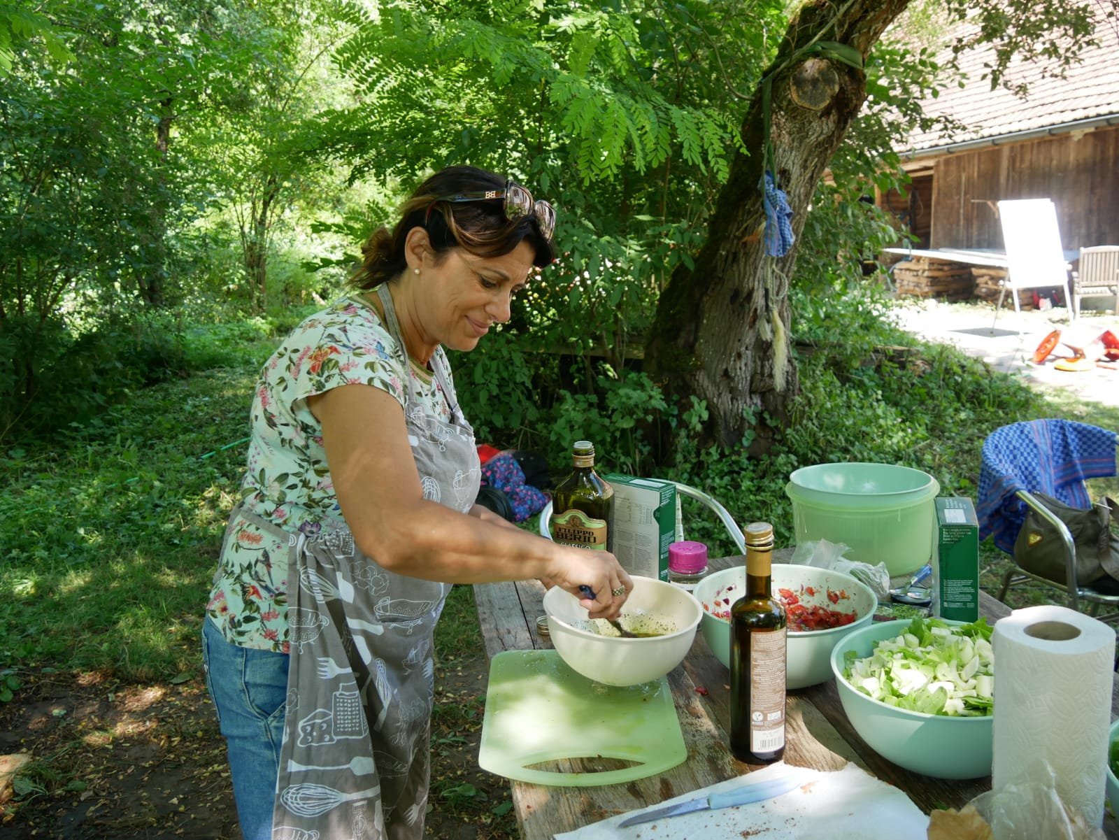 Frau im Freien, die an einem Holztisch unter Bäumen Salat und Zutaten zubereitet, umgeben von Schüsseln und Olivenöl.