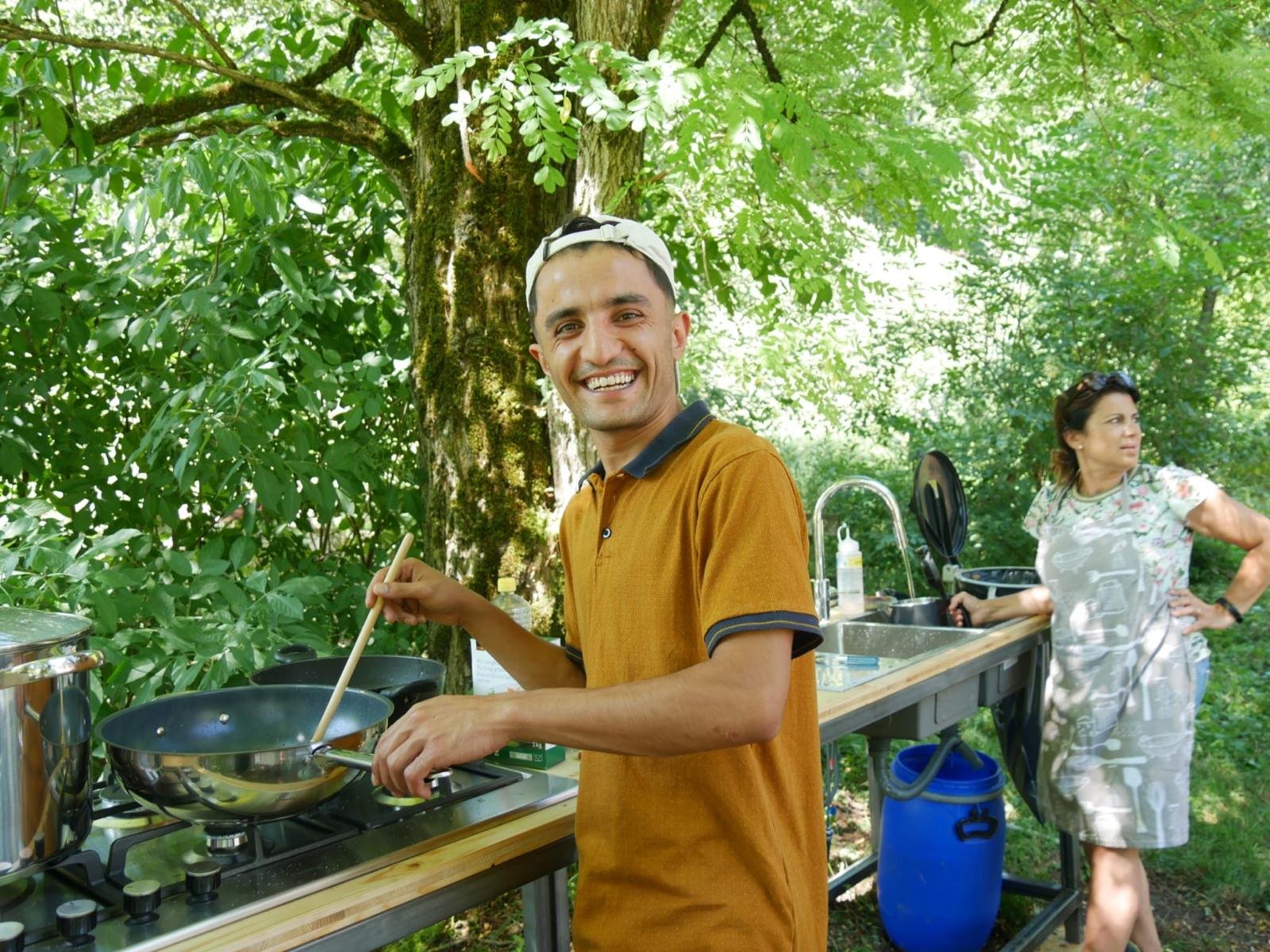 Zwei Personen in einer Outdoor-Küche, eine Person kocht an einem Herd unter einem Baum, die andere steht daneben. Im Hintergrund sind Bäume zu sehen. Sommertag.