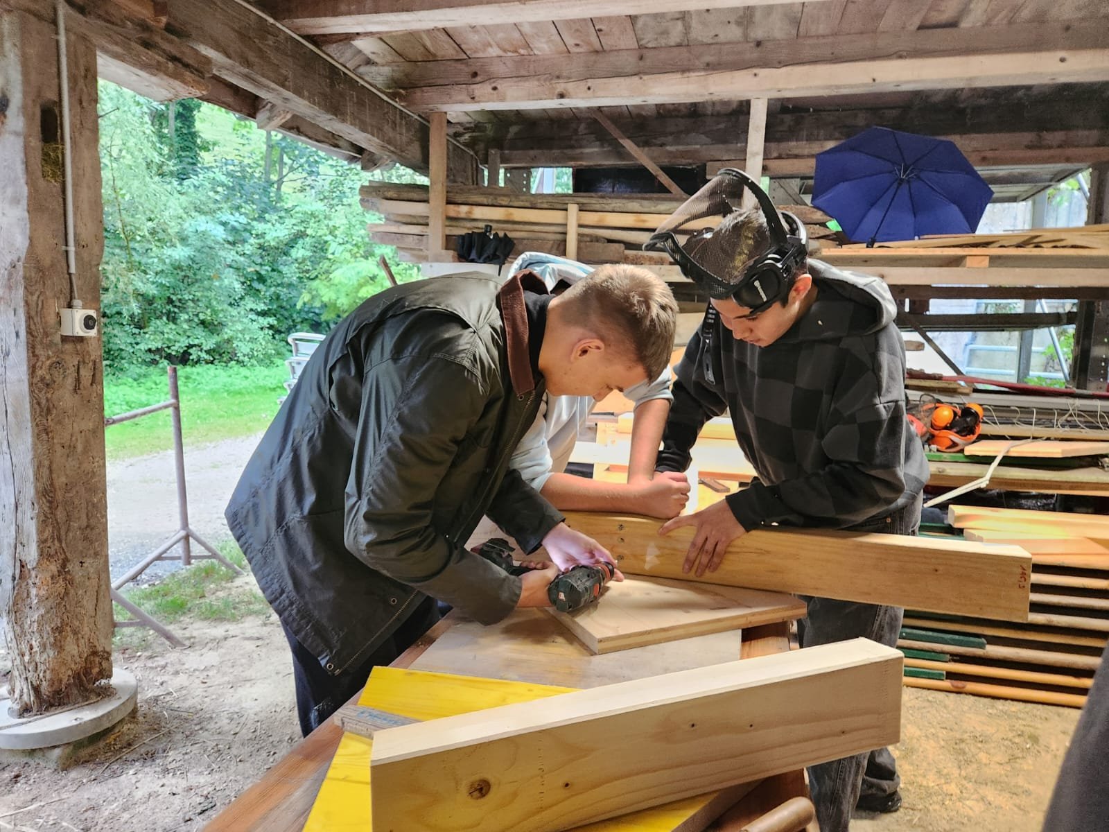 Zwei Personen arbeiten in einer Werkstatt mit Holz, verwenden Werkzeug auf einem Arbeitstisch, umgeben von Holzregalen und einem Waldhintergrund. Eine Person trägt einen Schutzhelm.