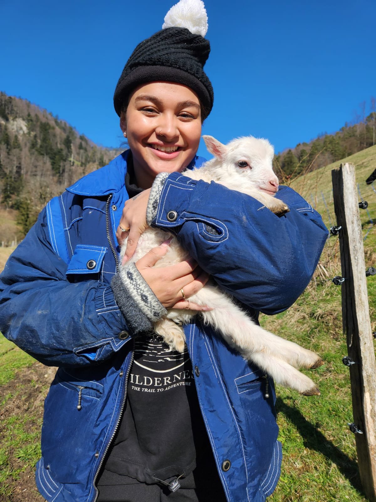 Eine Person in blauer Jacke und schwarzer Mütze hält ein kleines, weißes Lamm im Arm. Der Hintergrund zeigt eine Landschaft mit blauem Himmel und grünen Hügeln.
