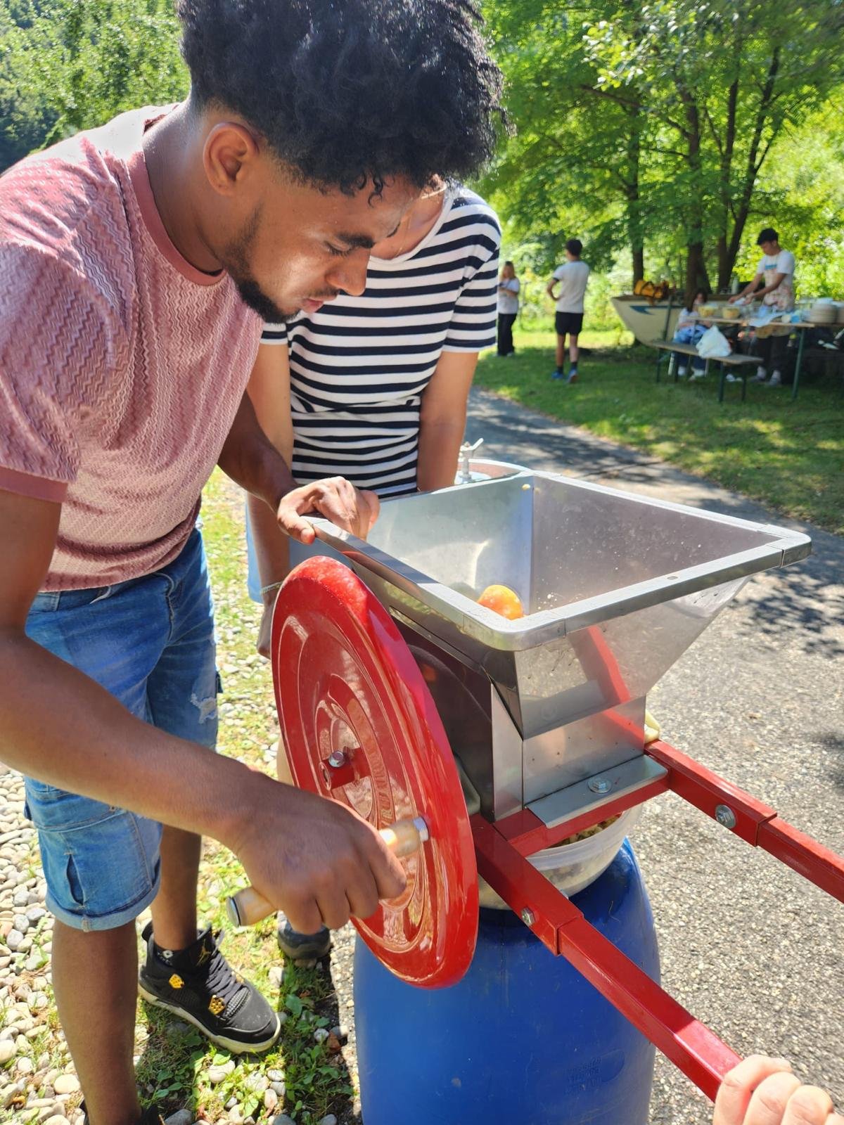 Zwei Personen bedienen eine manuelle Obstpresse im Freien. Eine Person dreht das rote Rad der Presse, während die andere zuschaut. Im Hintergrund sind Bäume und Menschen an einem Tisch zu sehen.