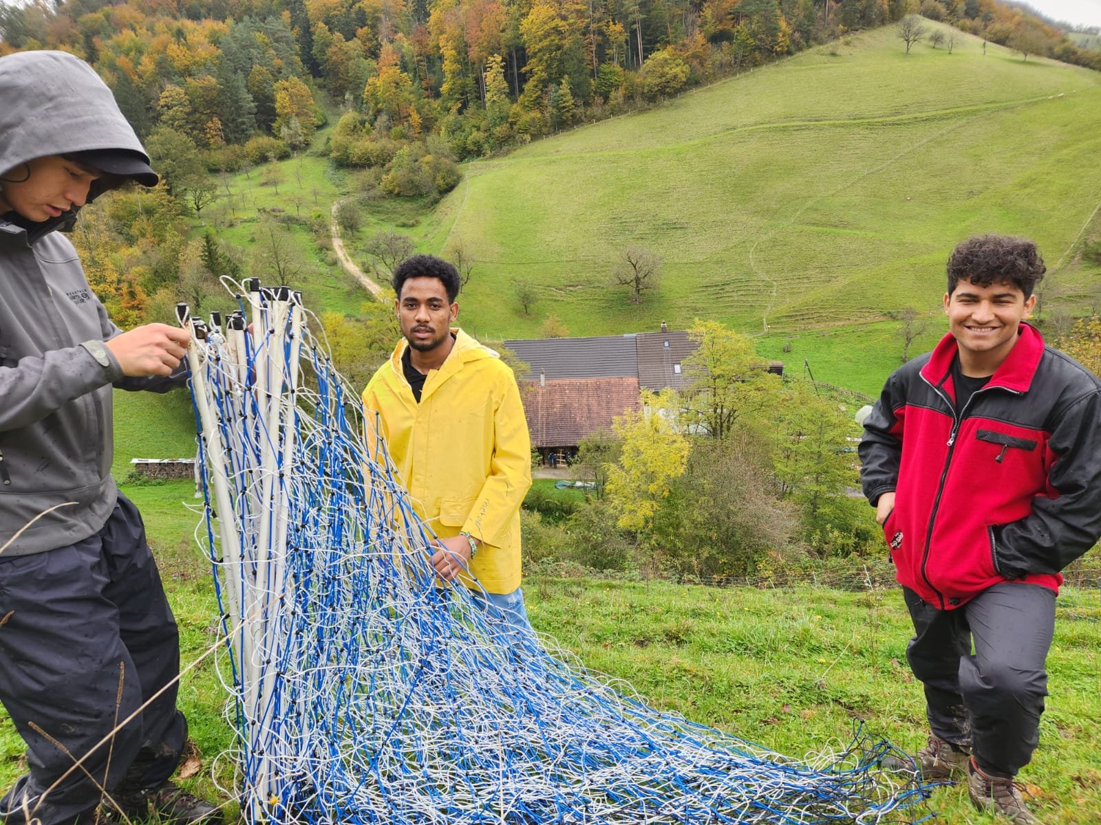 Drei Männer stehen auf einem Hügel mit grüner Landschaft im Hintergrund. Einer trägt einen grauen Regenmantel und untersucht ein blau-weißes Gitter oder Netze. Ein anderer Mann trägt einen gelben Regenmantel und schaut in die Kamera. Der dritte Mann,