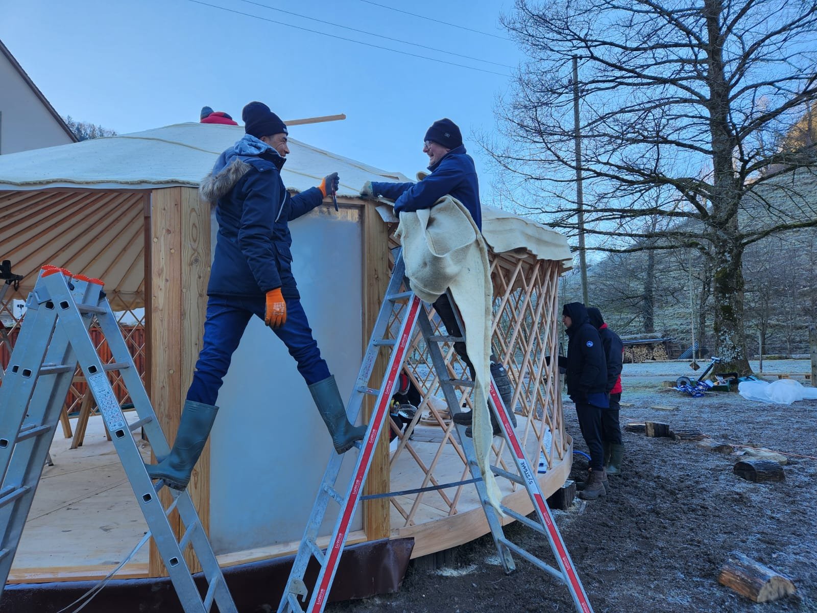 Personen bauen eine Jurte im Freien. Zwei Personen stehen auf Leitern und arbeiten am Dach, während andere auf dem Boden stehen. Die Jurte hat eine Holzstruktur und wird mit einer Plane bedeckt. Im Hintergrund sind Bäume zu sehen.
