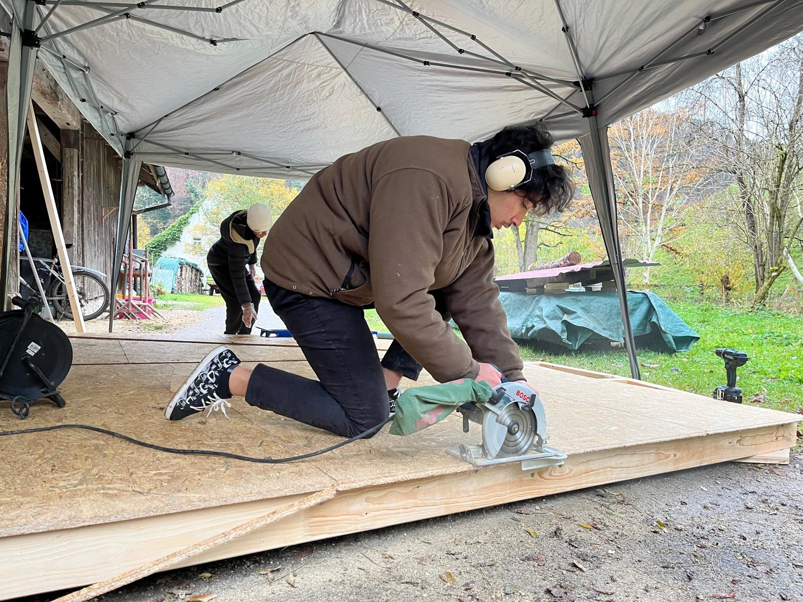 Zwei Personen arbeiten unter einem Zelt im Freien. Eine Person schneidet mit einer Kreissäge Holzplatten, die andere Person steht im Hintergrund. Sie tragen Gehörschutz. Der Arbeitsplatz befindet sich in einer ländlichen Umgebung.
