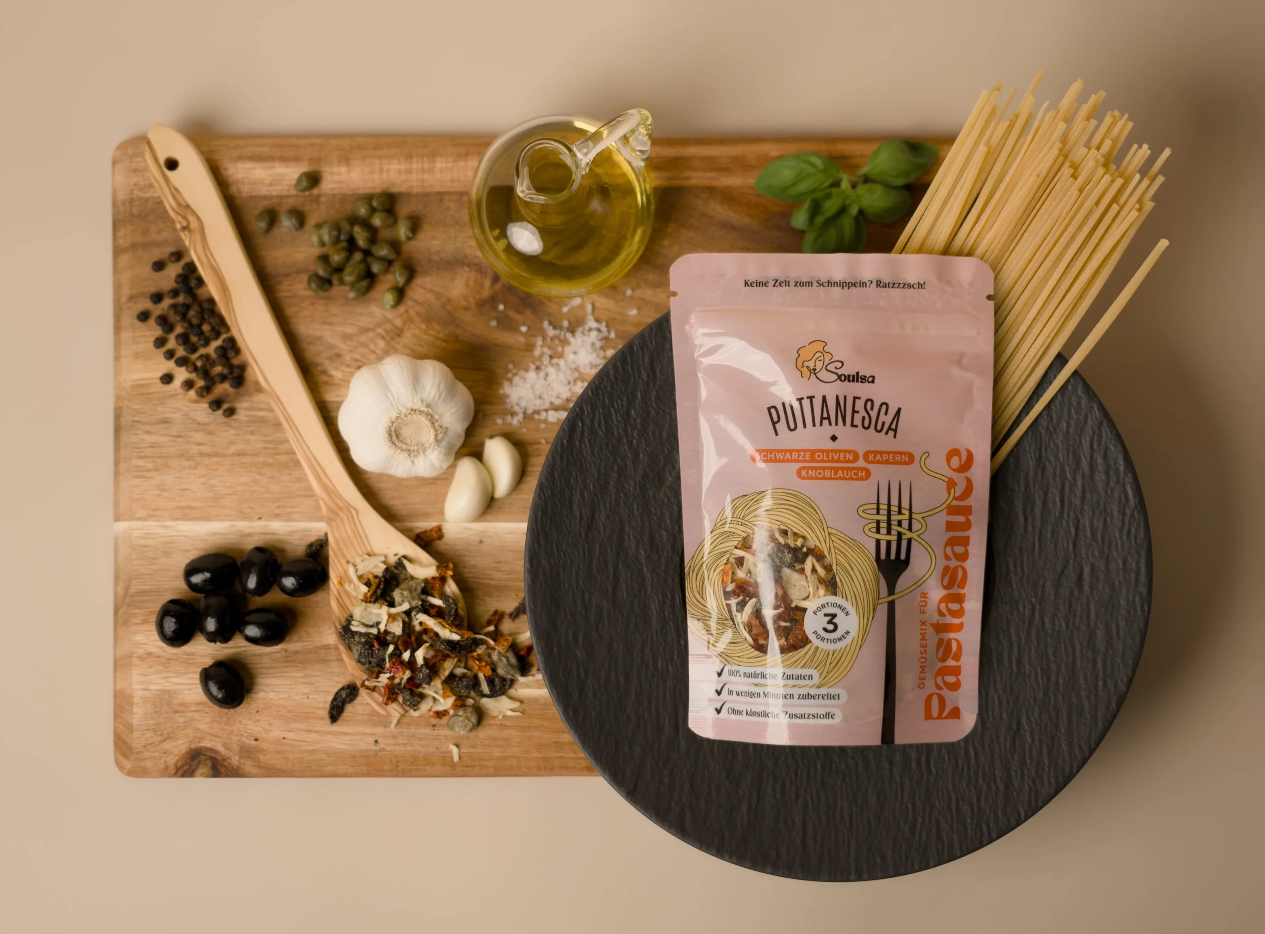 Holzbrett mit schwarzen und grünen Oliven, Knoblauch, Meersalz, Pfeffer, und weitere Zutaten, sowie eine Flasche Olivenöl, Basilikum, und frisch verpackte Puttanesca Pasta.