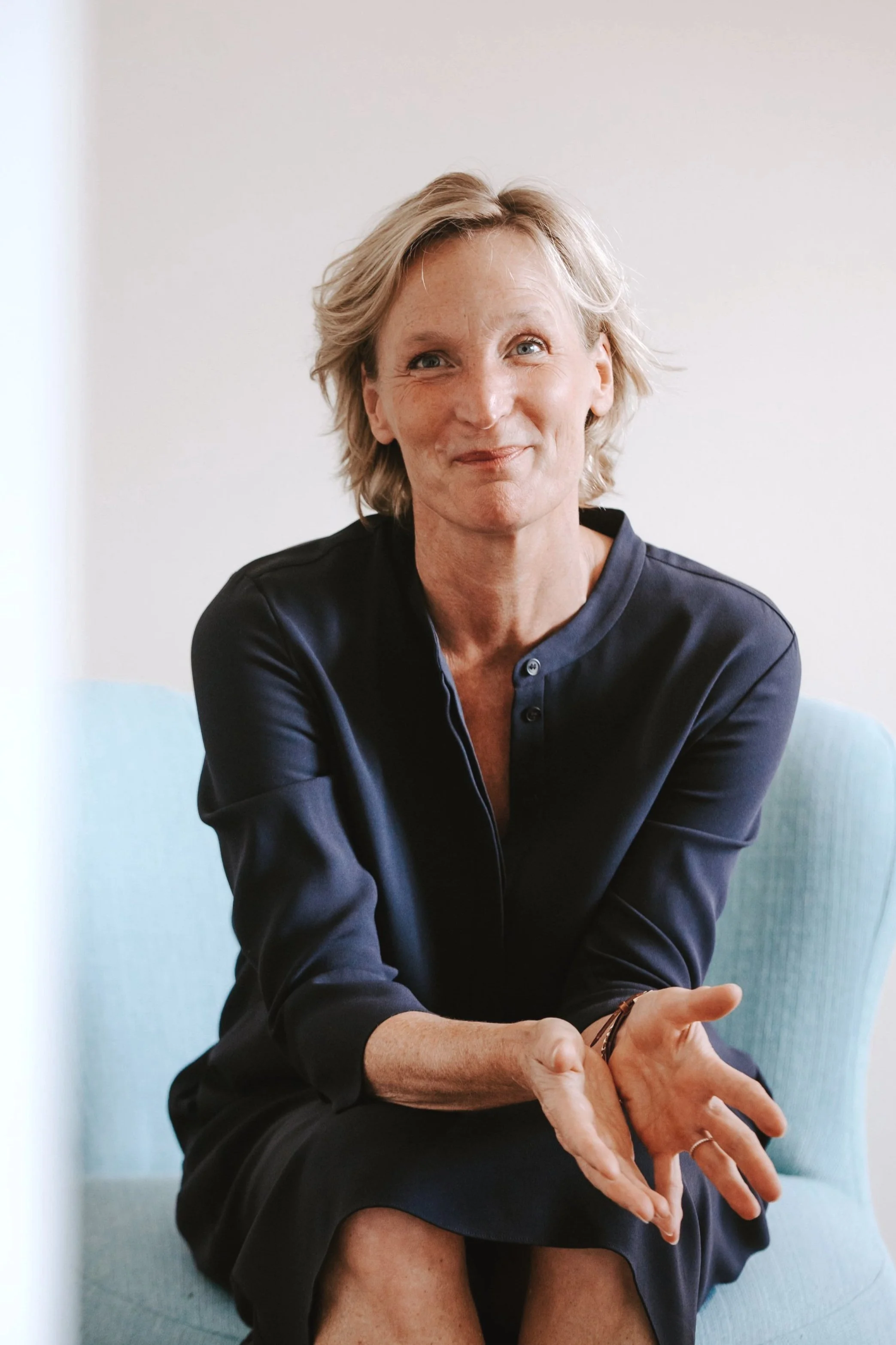 A woman with short blonde hair, wearing a navy blue dress, sitting on a light blue chair, smiling and gesturing with her hands.