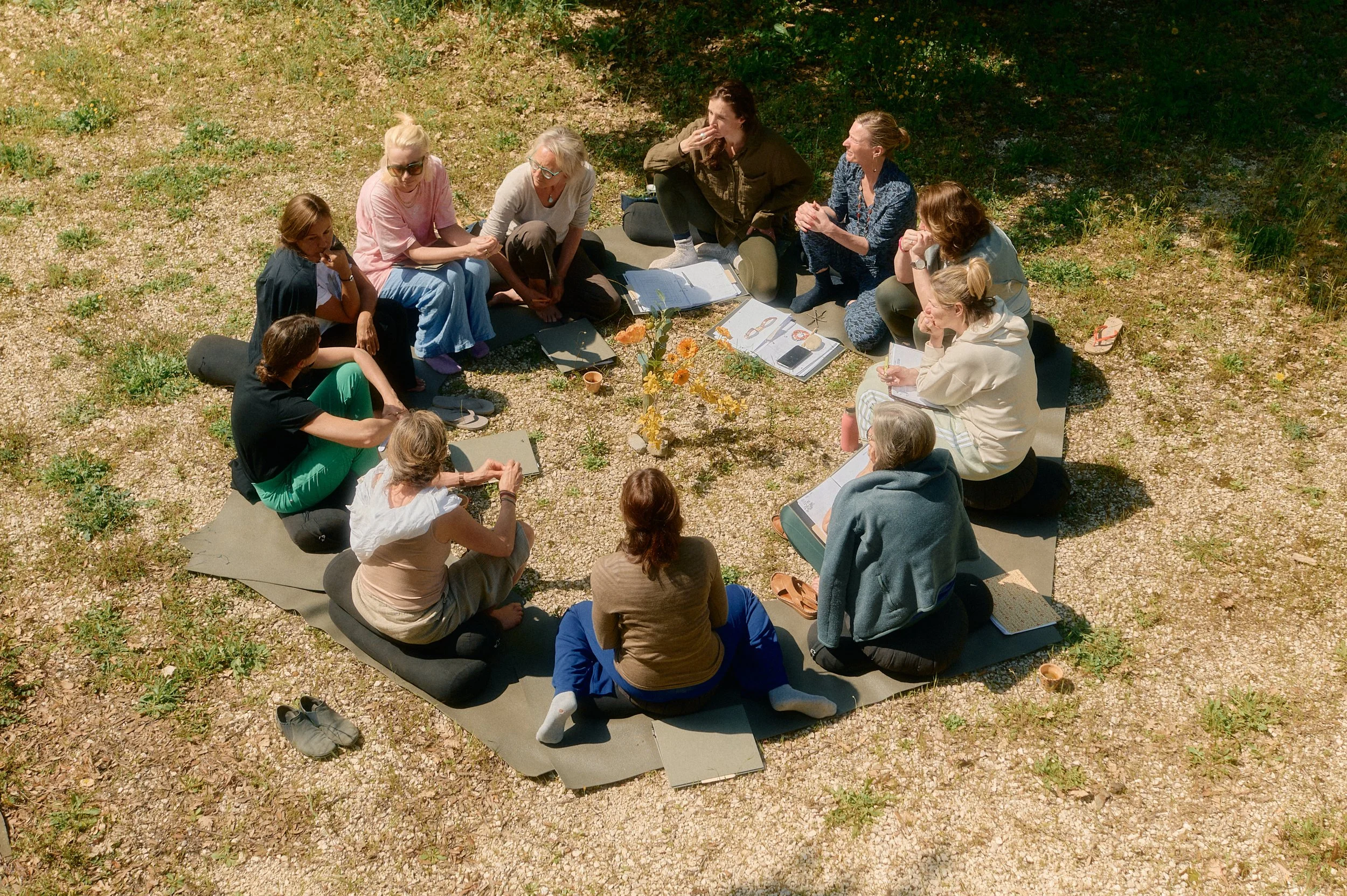 A group of twelve people sitting in a circle on mats outdoors on a gravel surface, engaging in a discussion with notebooks and a flower vase in the center.