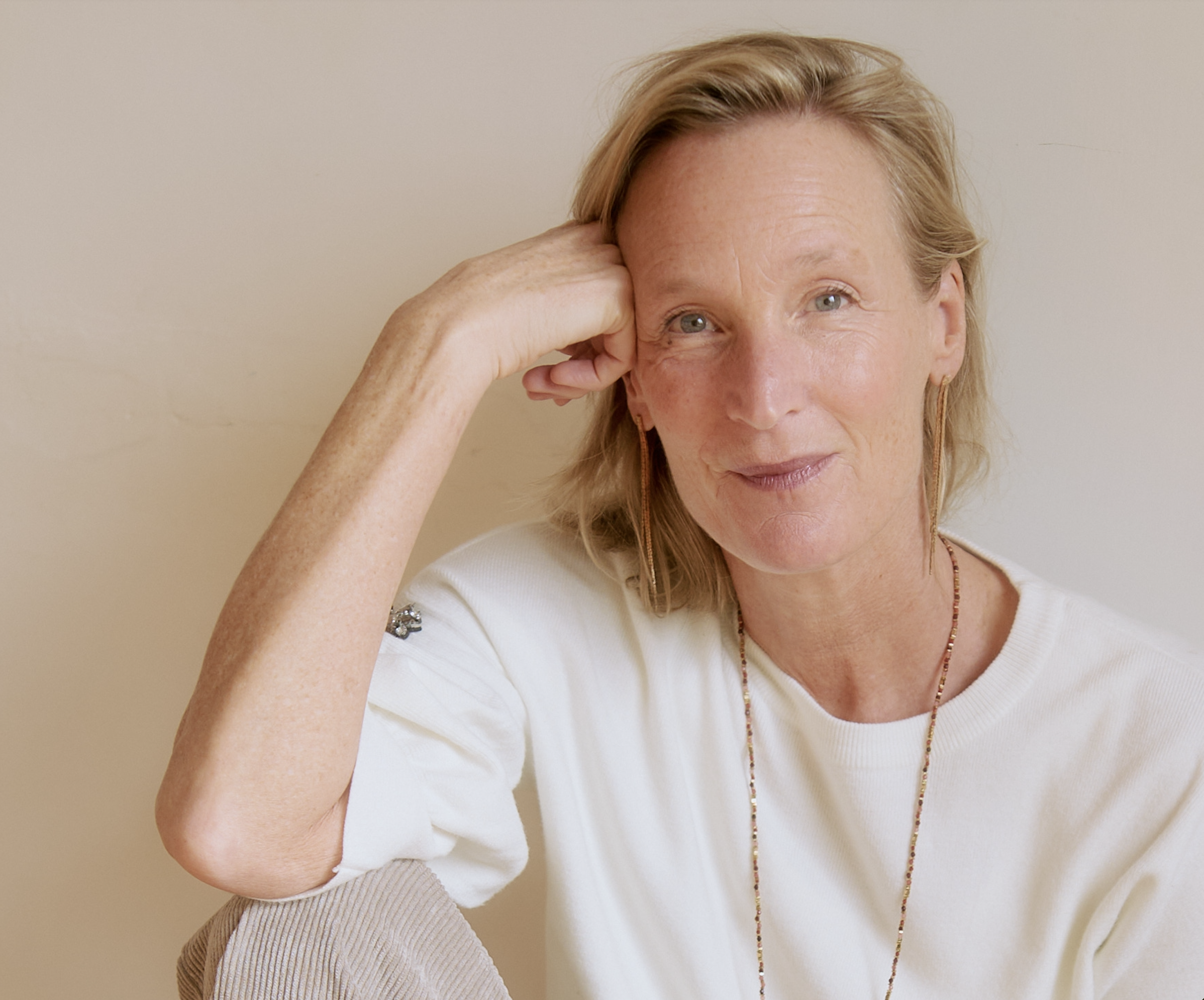 A woman with blonde hair, wearing a white top and jewelry, resting her head on her hand and smiling at the camera against a neutral background.