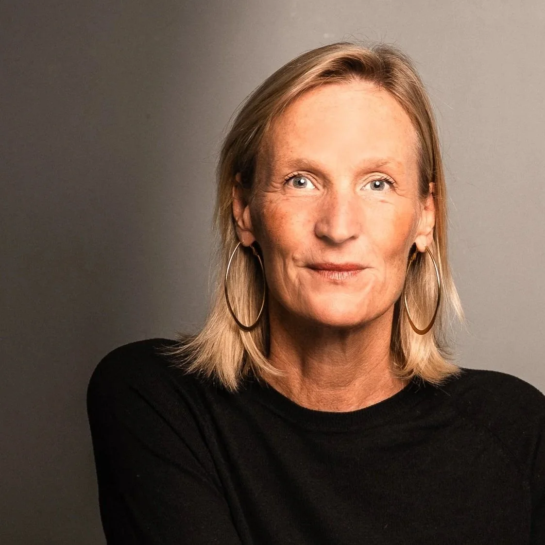 A middle-aged woman with blonde hair, blue eyes, and hoop earrings, wearing a black top, against a plain gray background.