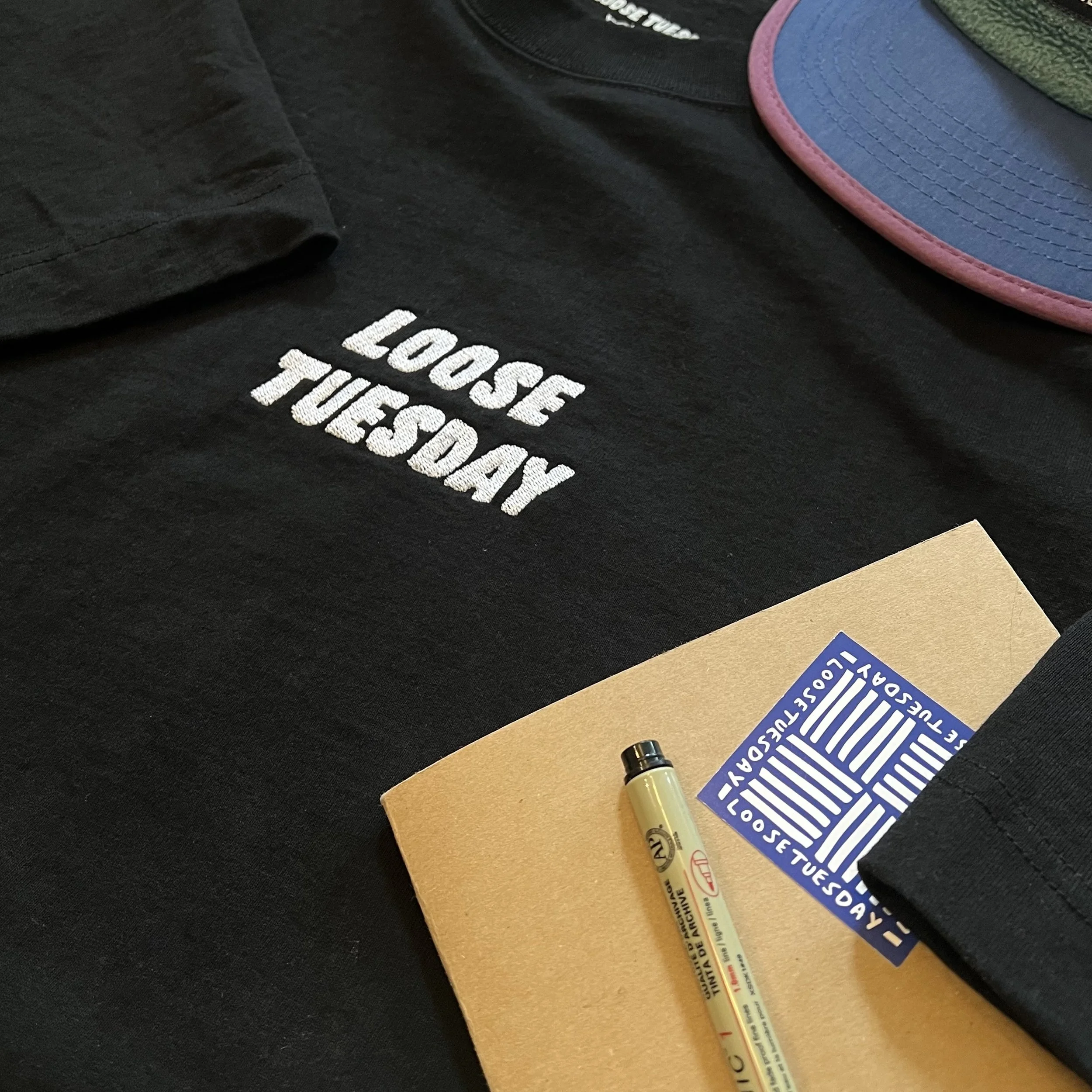 Loose Tuesday Everyday Tee — chest embroidery detail on black, styled with accessories