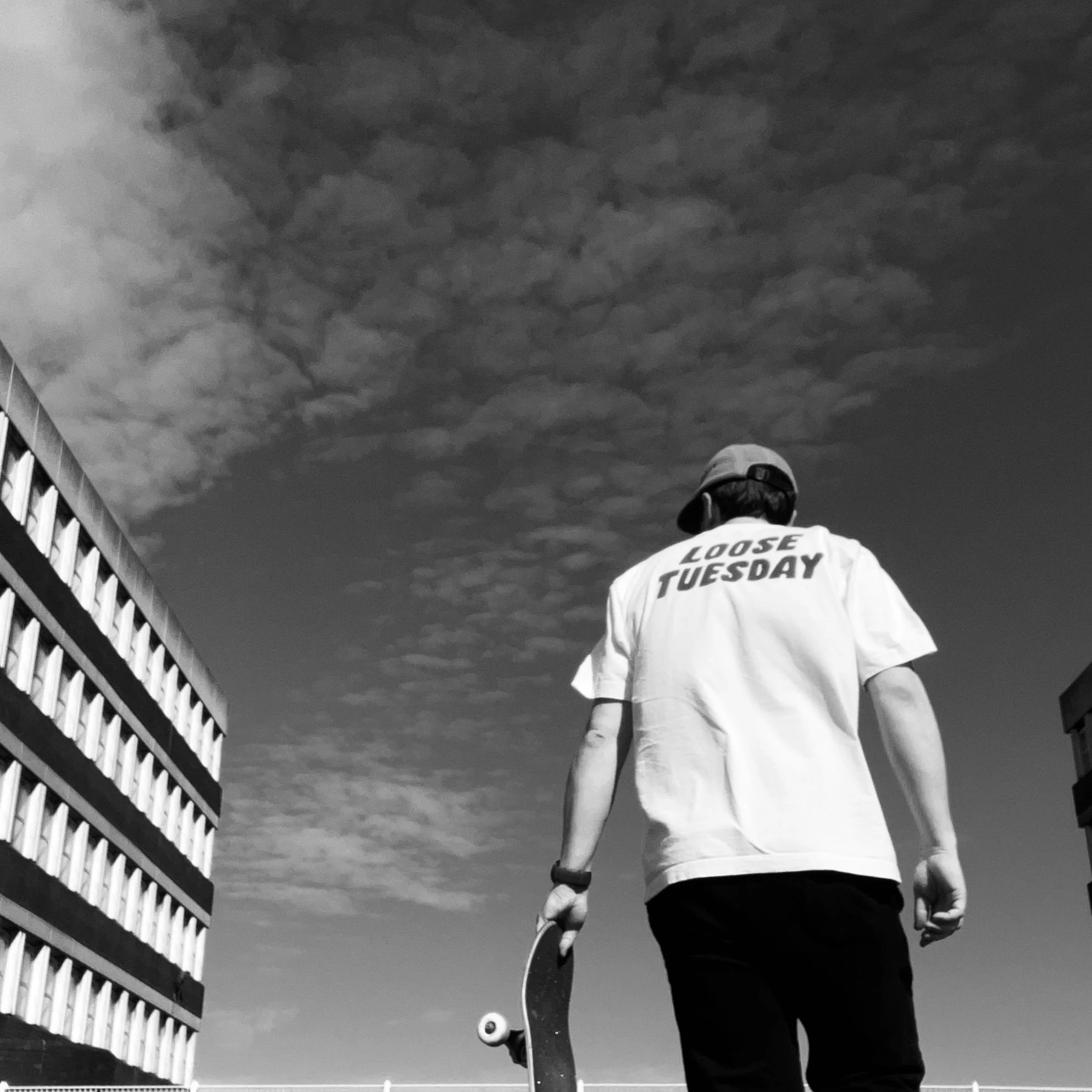 Person holding a skateboard, shot from behind, wearing a white Loose Tuesday graphic tee