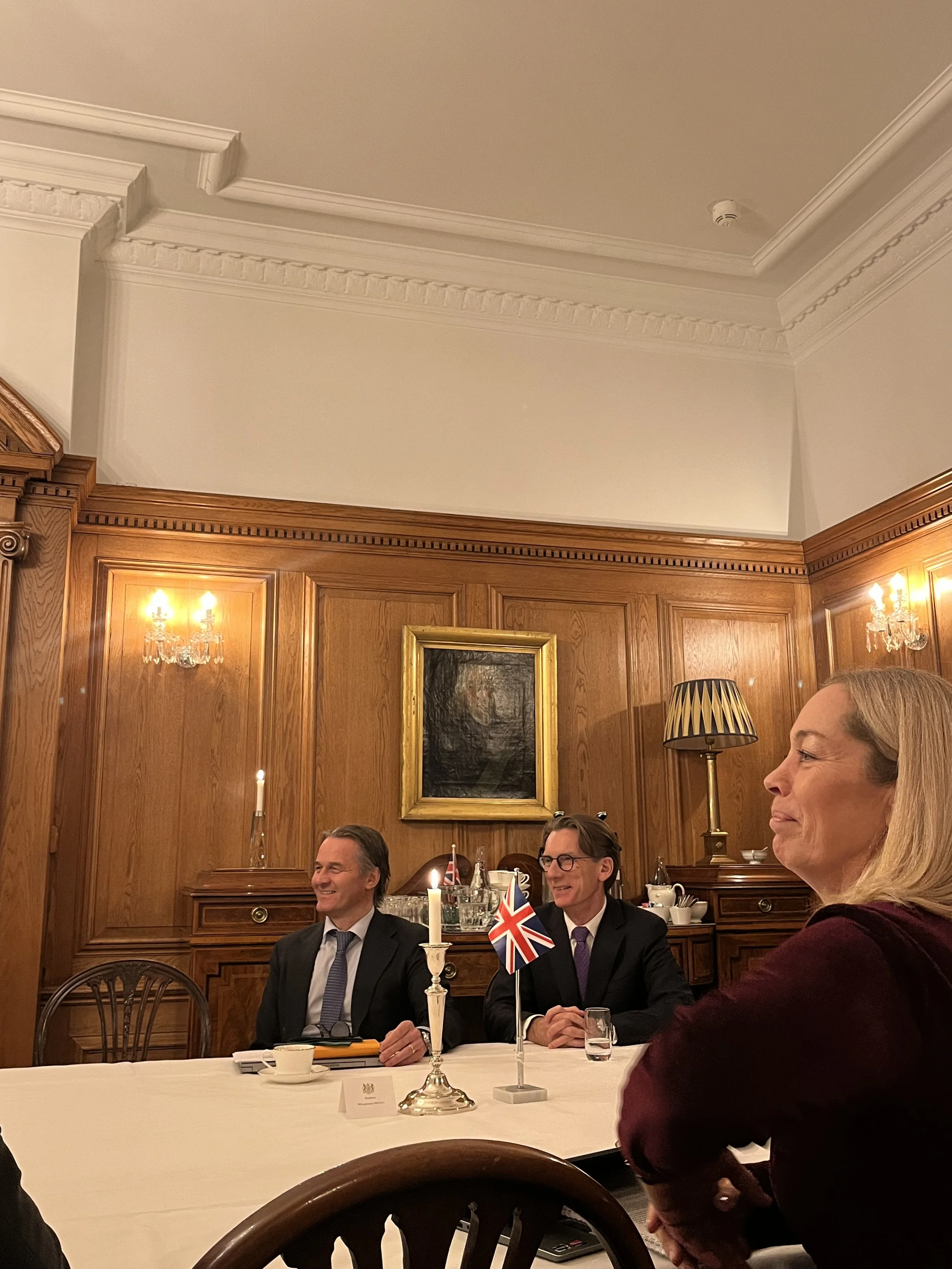 A formal meeting with two men in suits and a woman sitting at a table in a wood-paneled room with candles and a British flag.