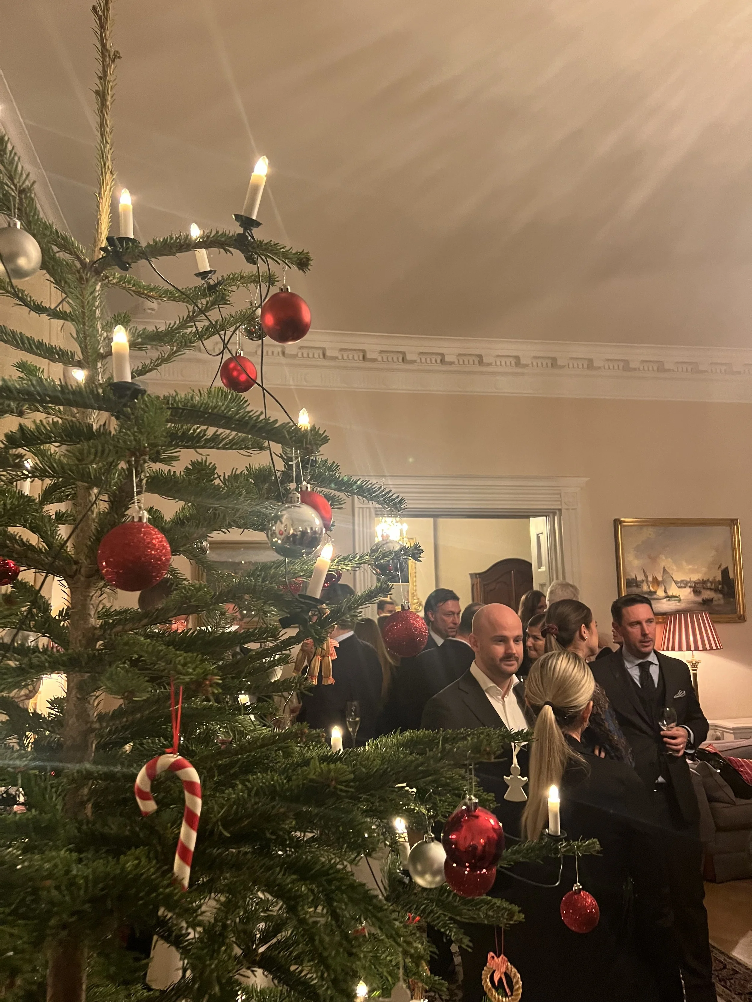 People gathered at a Christmas party with a decorated Christmas tree in the foreground and a cozy living room in the background.