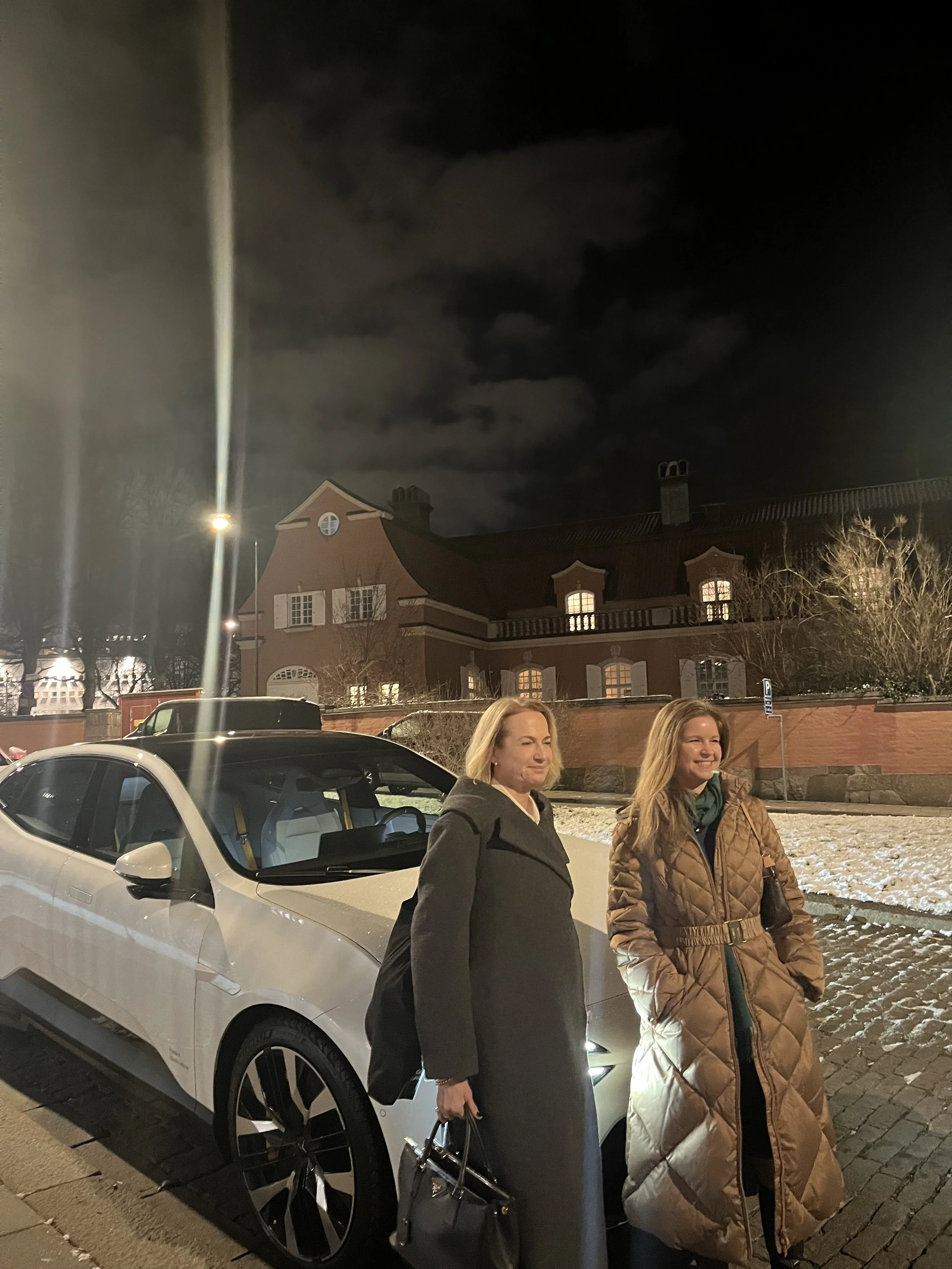 Two women standing next to a white car on a cobblestone street at night, with a large house and dark cloudy sky in the background.