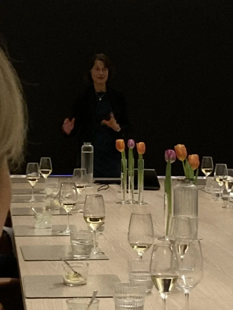 Woman giving a presentation in a dimly lit room with a long table set with wine glasses, water glasses, small candles, and vases with colorful tulips.