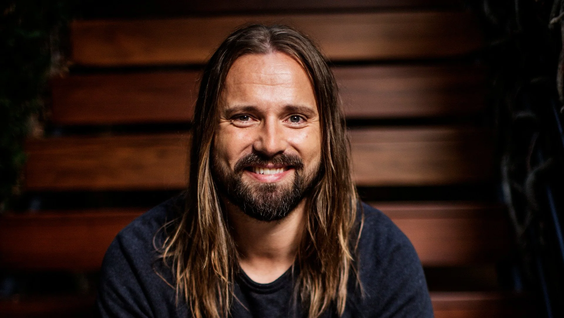 Max Martin, Music producer. Man with long hair and beard smiling, sitting on wooden stairs, wearing a dark shirt.