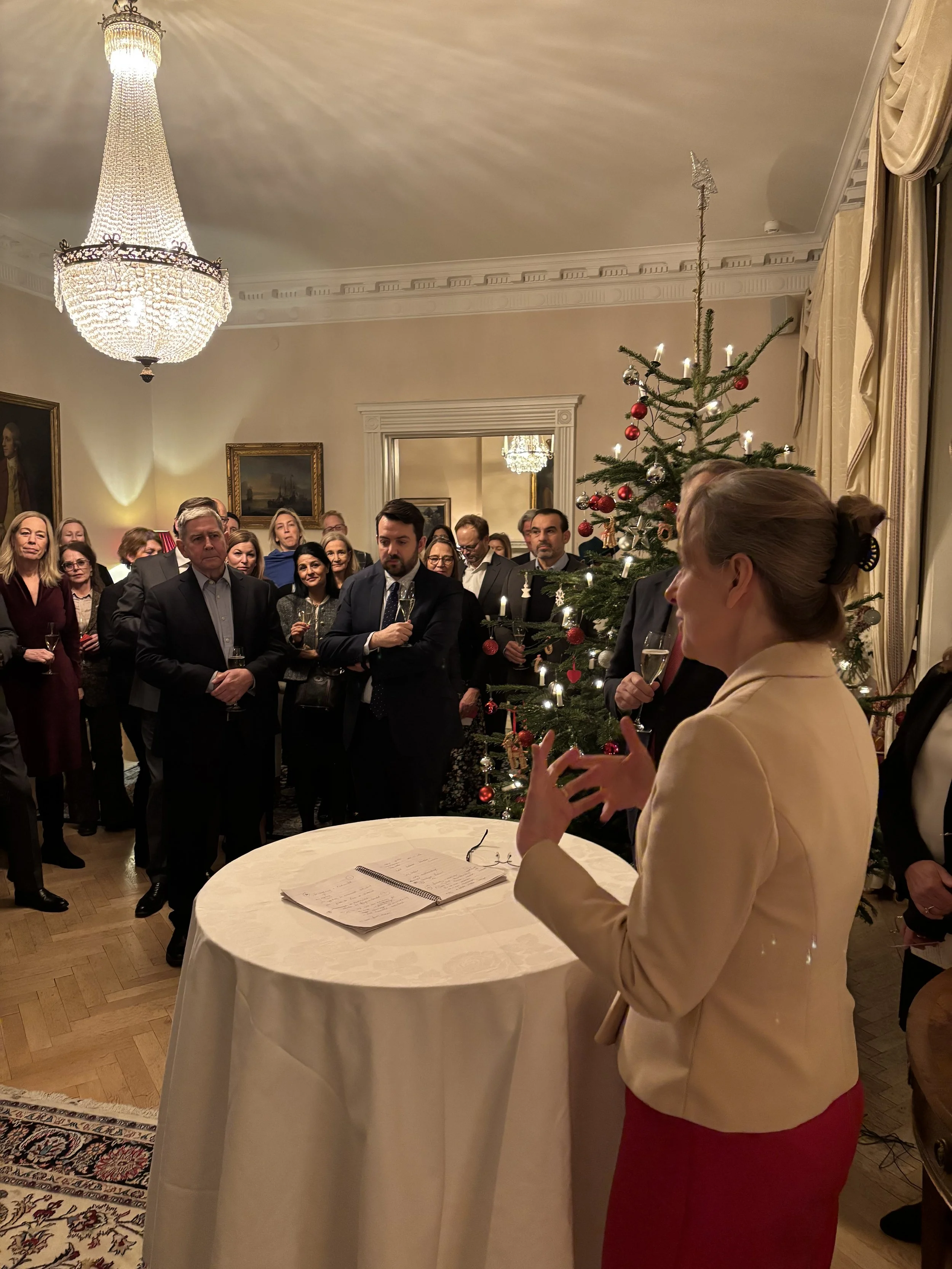 A woman giving a speech at a Christmas gathering in a decorated room with a Christmas tree, chandelier, and portrait paintings, as guests listen.