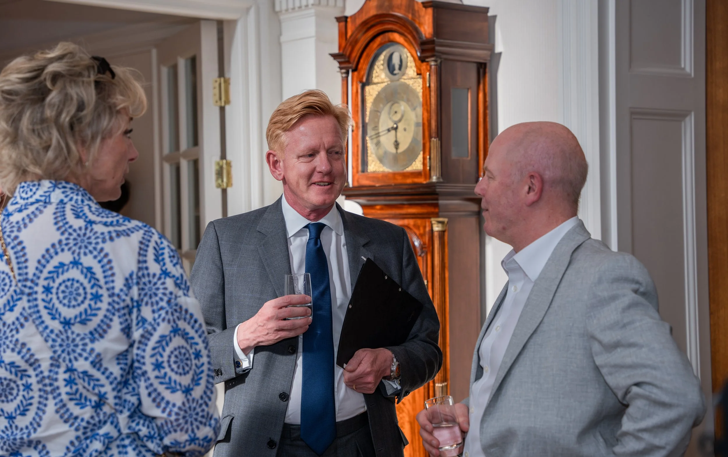 Three people having a conversation at a social event in a room with a grandfather clock in the background. They are dressed in business casual attire and holding drinks.