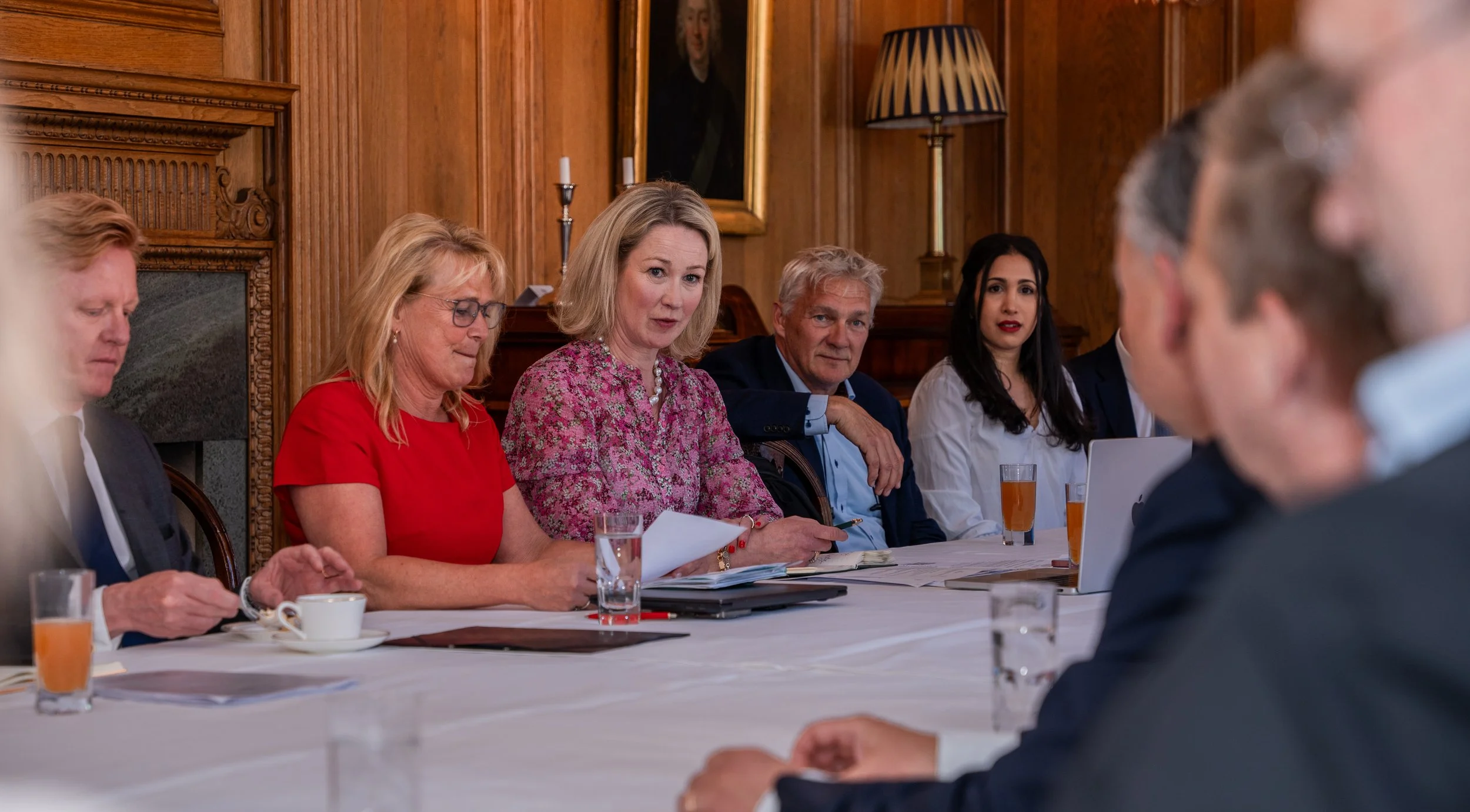 A group of people sitting at a long, white table in a wood-paneled room, engaged in a meeting or discussion, with one woman speaking and others listening, some with drinks and laptops.
