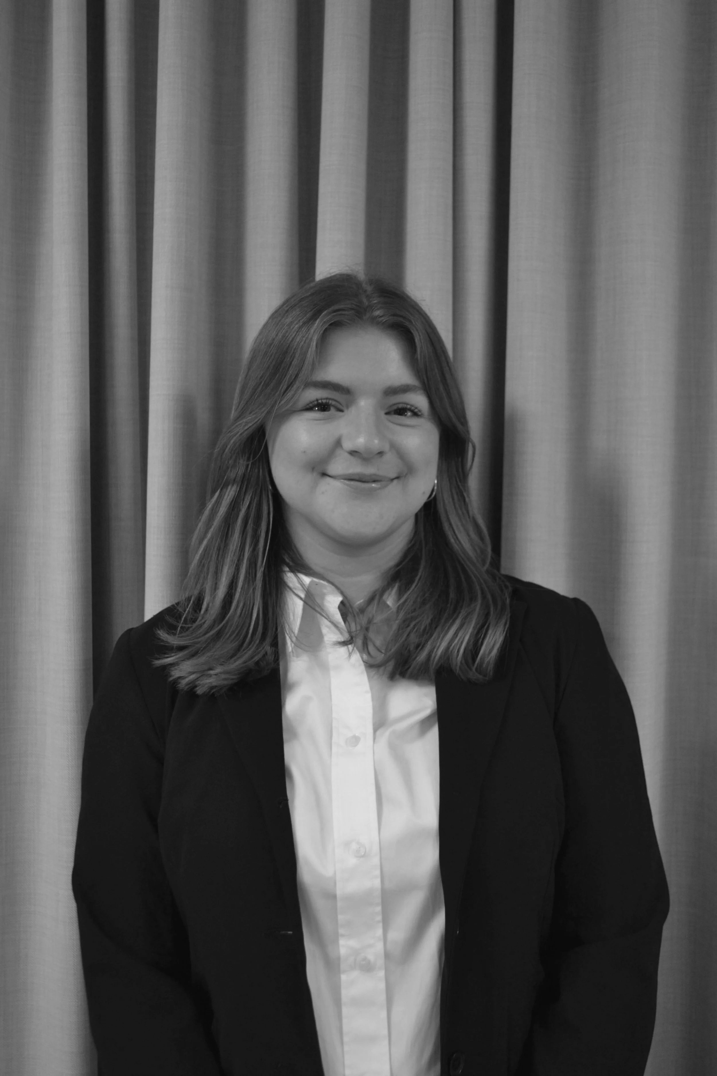 A young man in a suit and tie standing next to a window, looking at the camera with a slight smile in black and white.