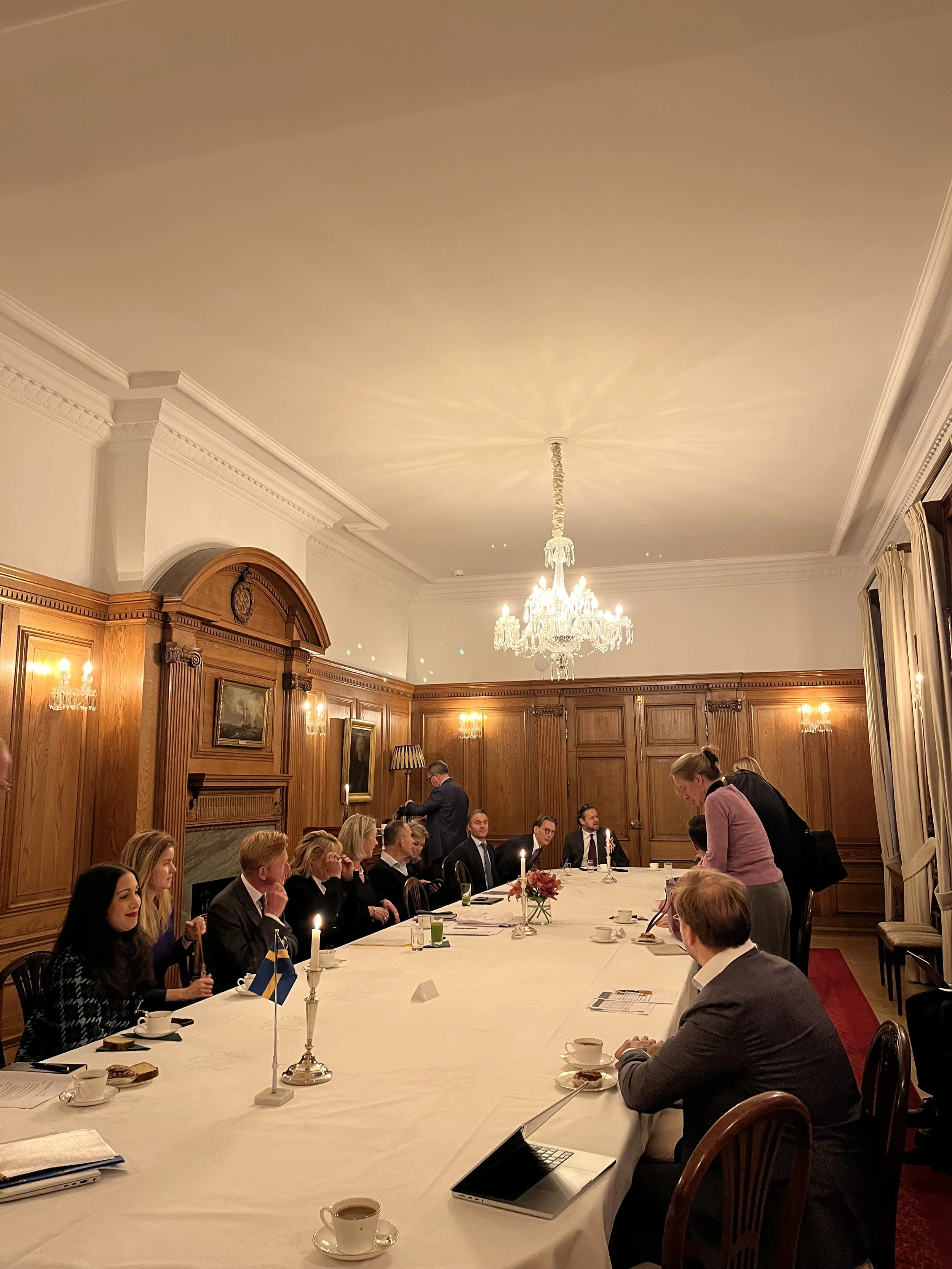 A formal meeting or dinner with multiple people seated around a long table in an elegant wood-paneled room with a chandelier, candles, and framed paintings.