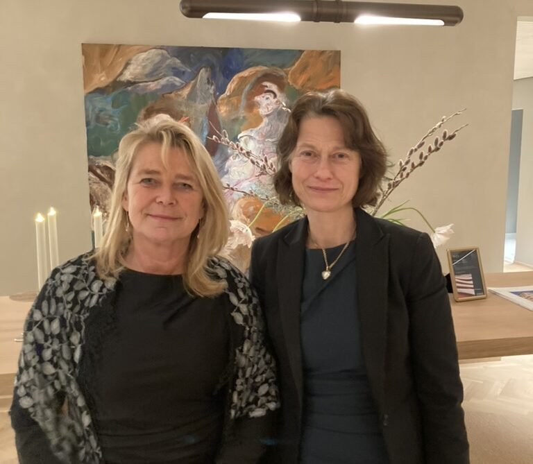 Two women standing side by side in an art gallery or similar setting, with a colorful abstract painting behind them and a small table with lit candles to the left.