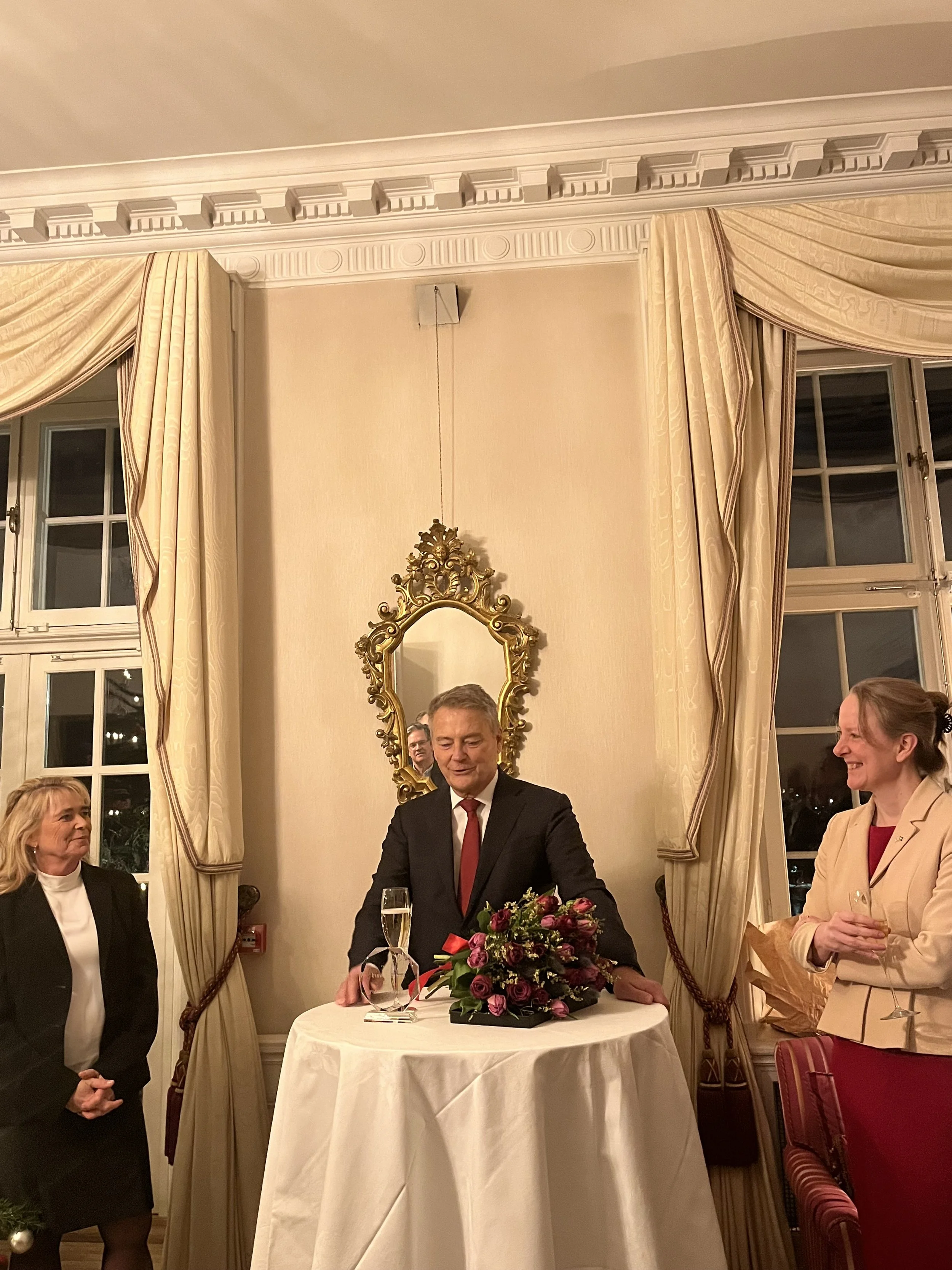 An indoor social gathering in an elegant room with cream-colored walls and large windows with draped curtains. A man in a dark suit and red tie stands behind a round table with a floral arrangement and a glass of champagne. Two women, one on each sid