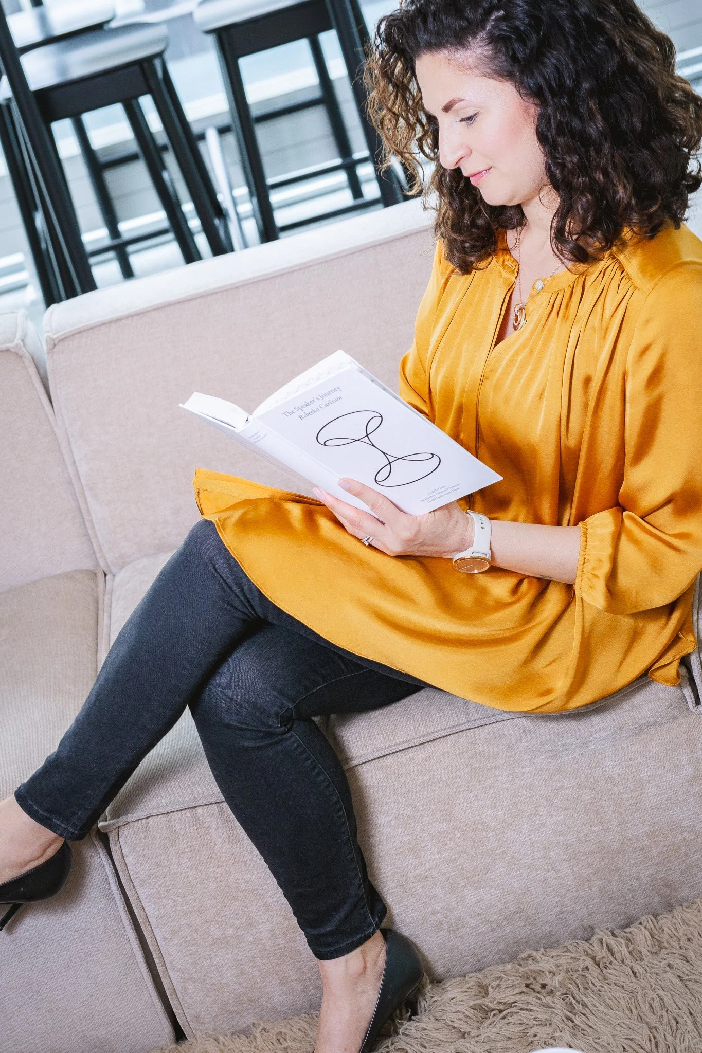 A woman with curly hair wearing a yellow blouse and black jeans, sitting on a beige sofa, reading a book in a bright room with barstools visible in the background.