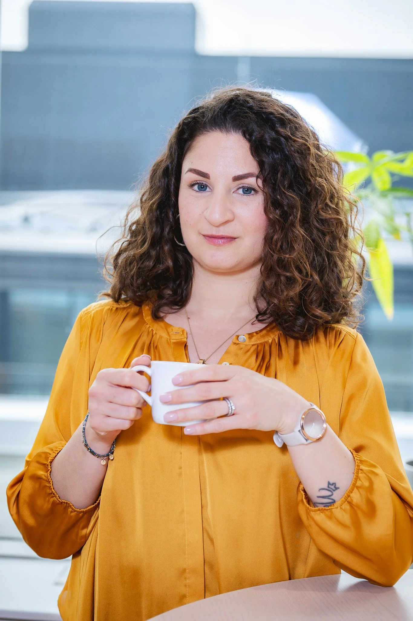 A woman with curly brown hair and blue eyes holding a white mug, wearing a mustard yellow blouse, sitting indoors near a window with a gray background and green plant to the side.