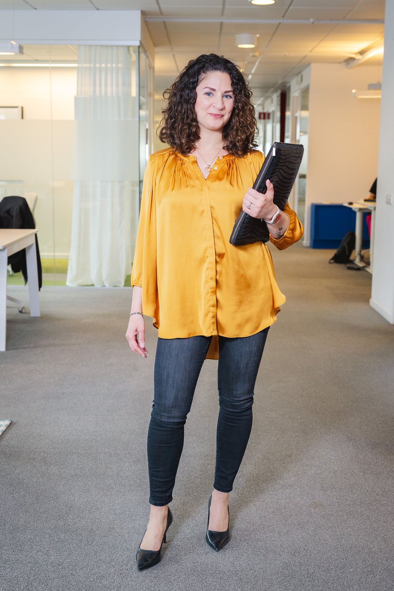 A woman with curly brown hair, wearing a yellow blouse and dark jeans, is standing in an office with carpeted flooring, holding a black folder, and wearing black high heels.