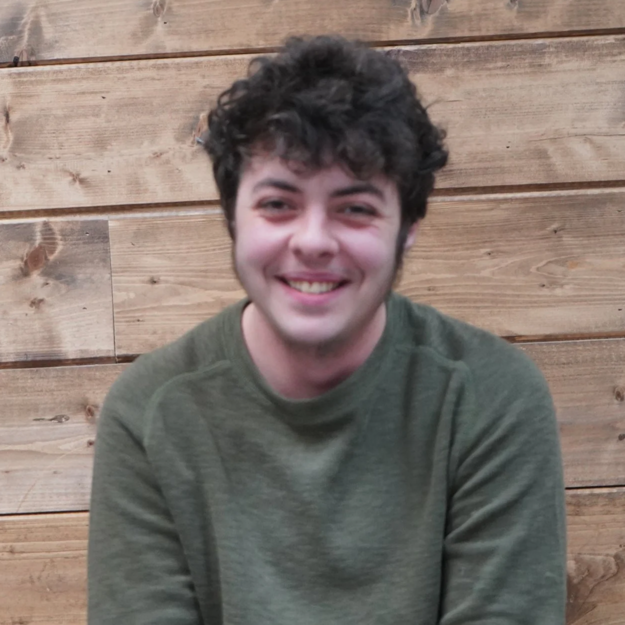 Young man with curly dark hair smiling, wearing a green sweater, seated against a wooden wall.