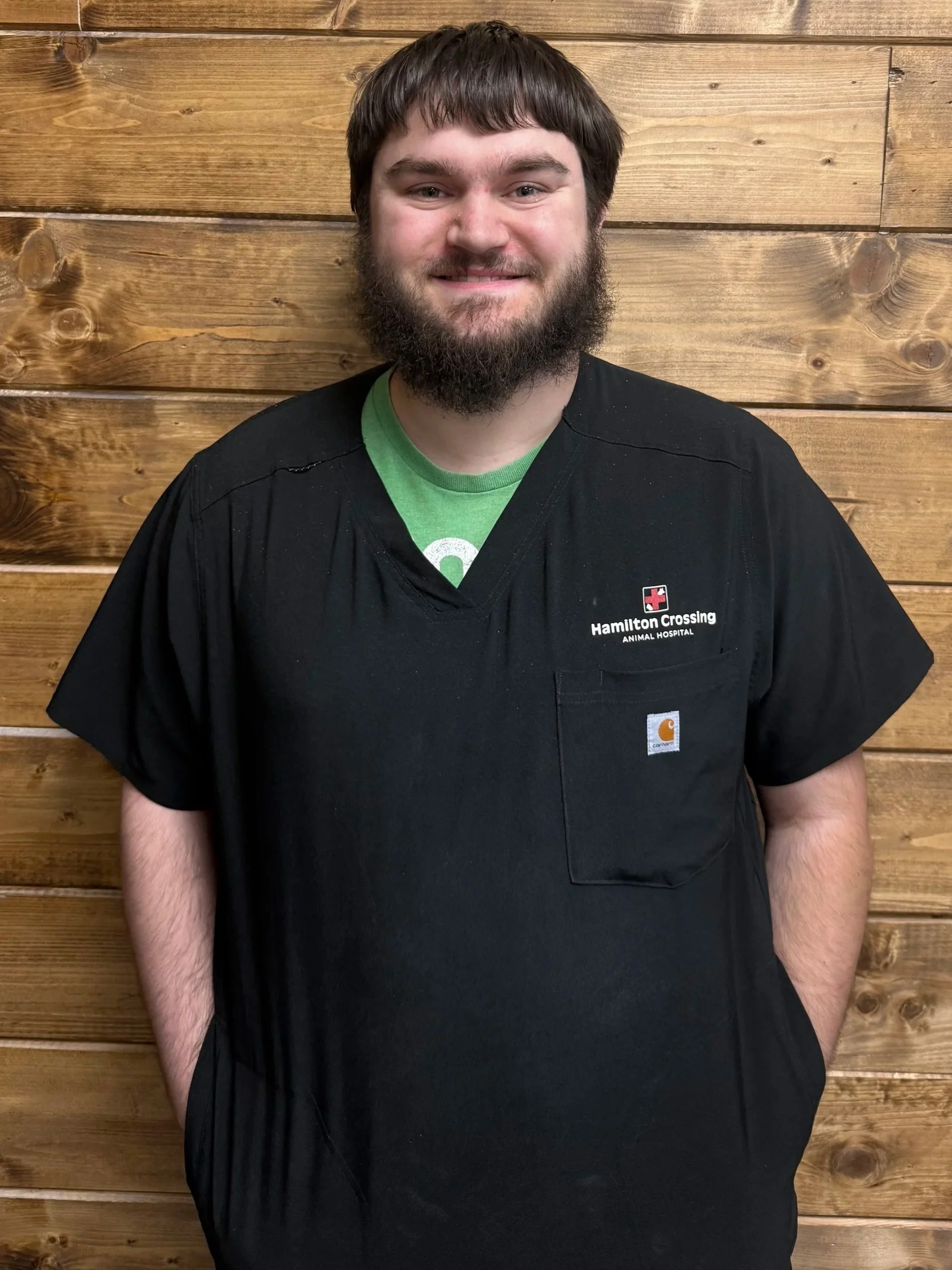 A man with a beard and short brown hair smiling, wearing a red and black plaid shirt, standing in front of a wooden wall.