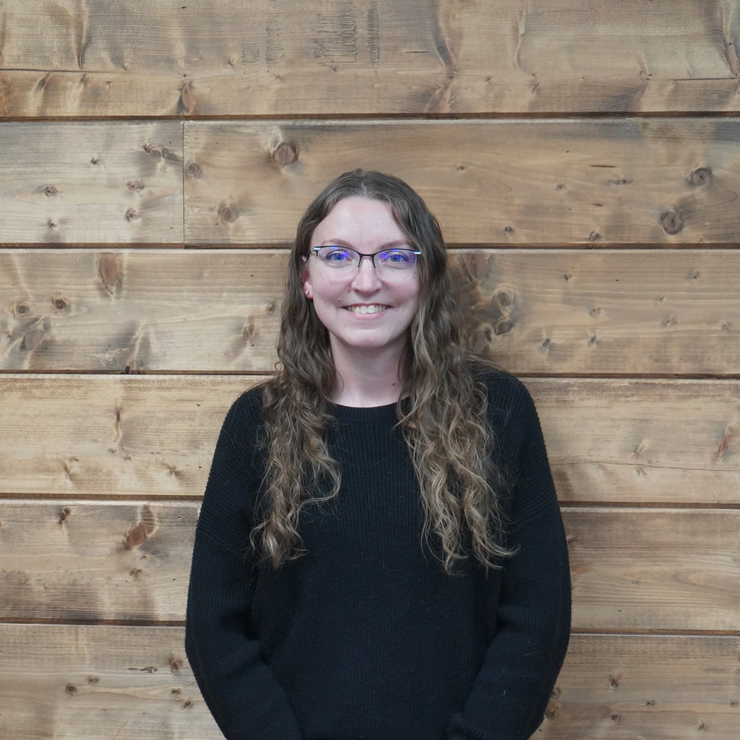 A woman standing in front of a wooden wall, wearing glasses, a black sweater, and jeans, smiling at the camera.