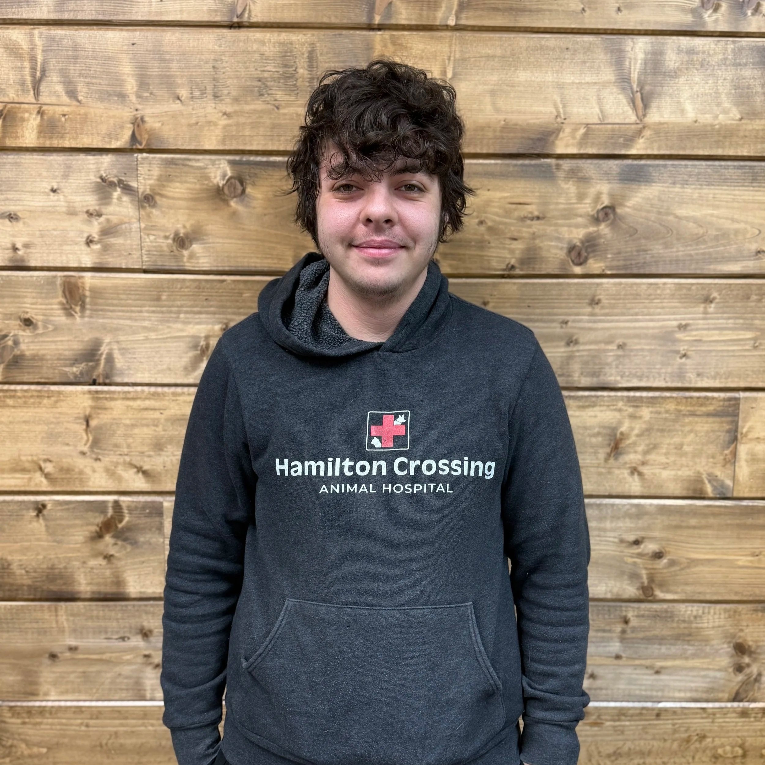 Young man with curly dark hair smiling, wearing a green sweater, seated against a wooden wall.